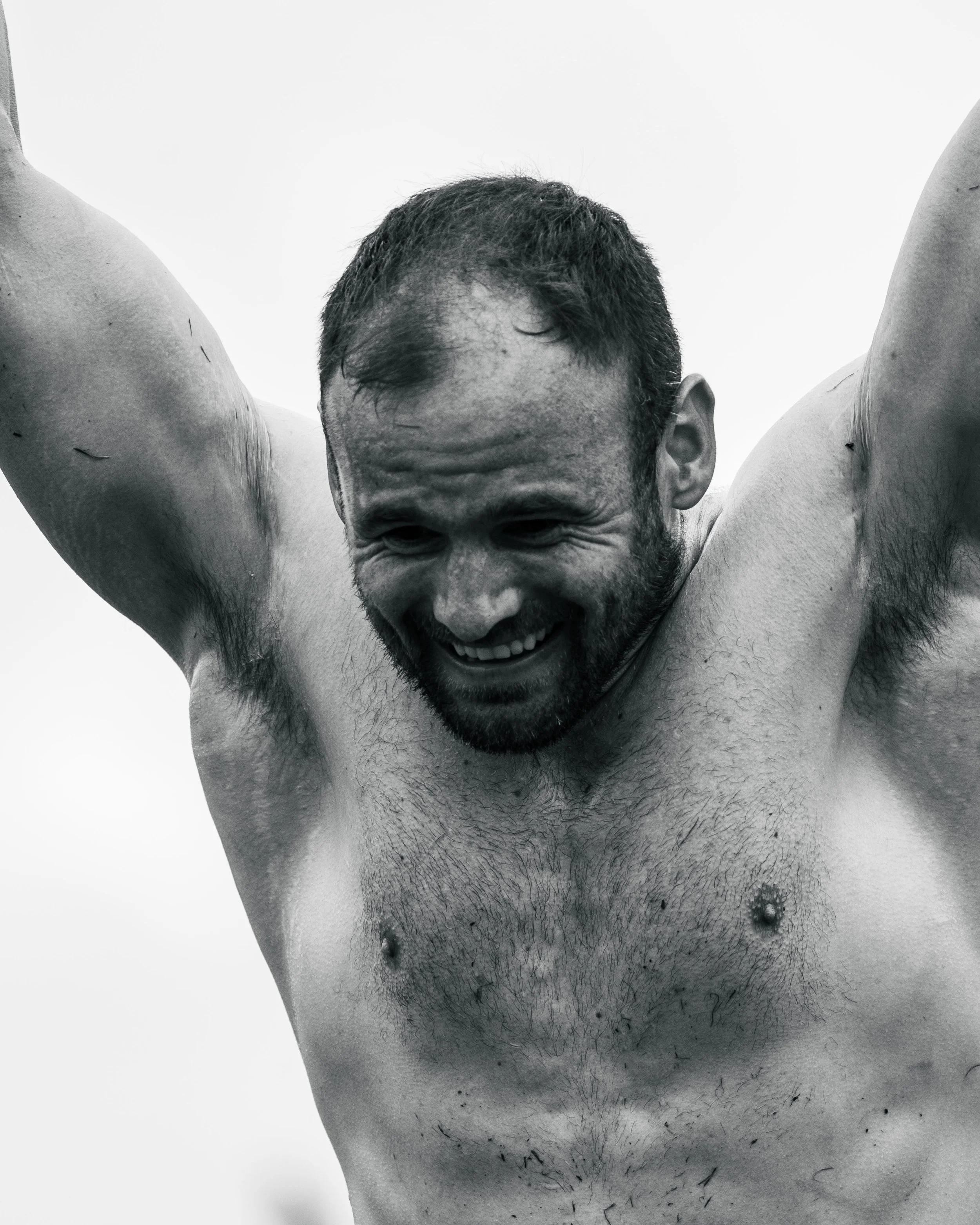 A shirtless man with a beard smiling and raising his arms.