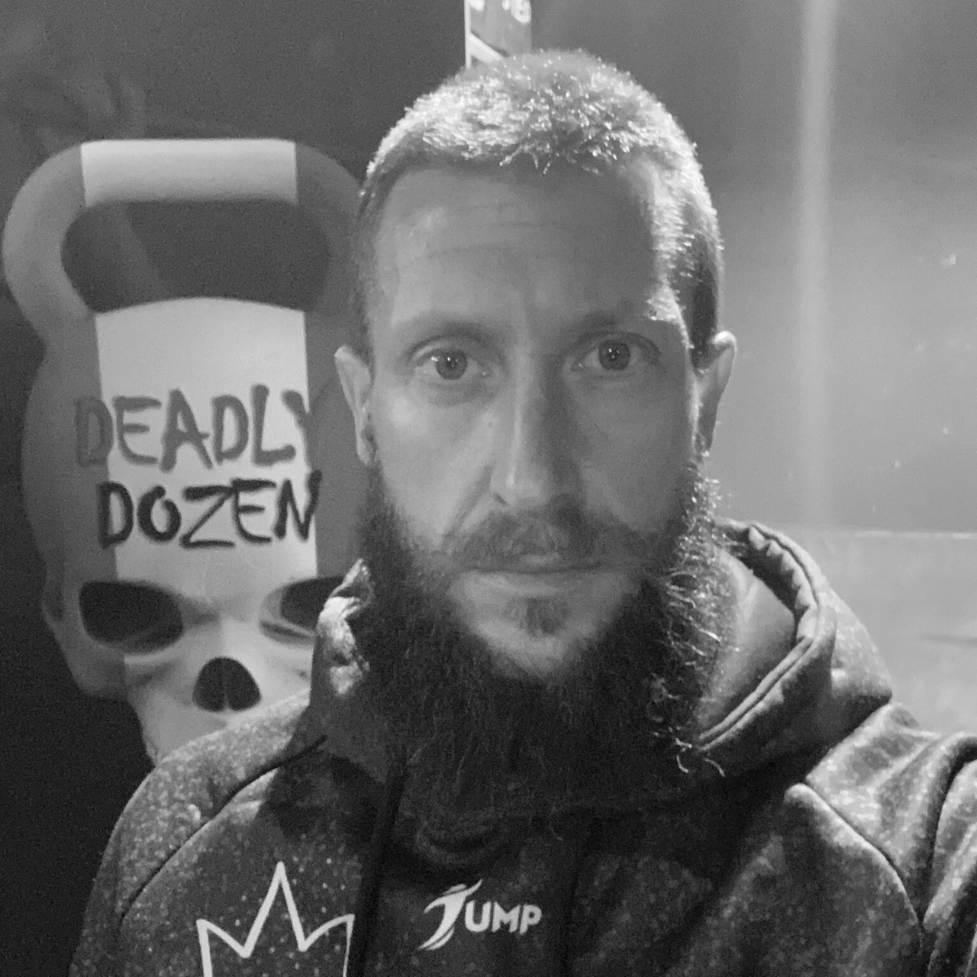 A man with a beard and short hair taking a selfie in front of a poster for the video game Deadly Dozen, which features a skull and the game's title. The photo is in black and white.