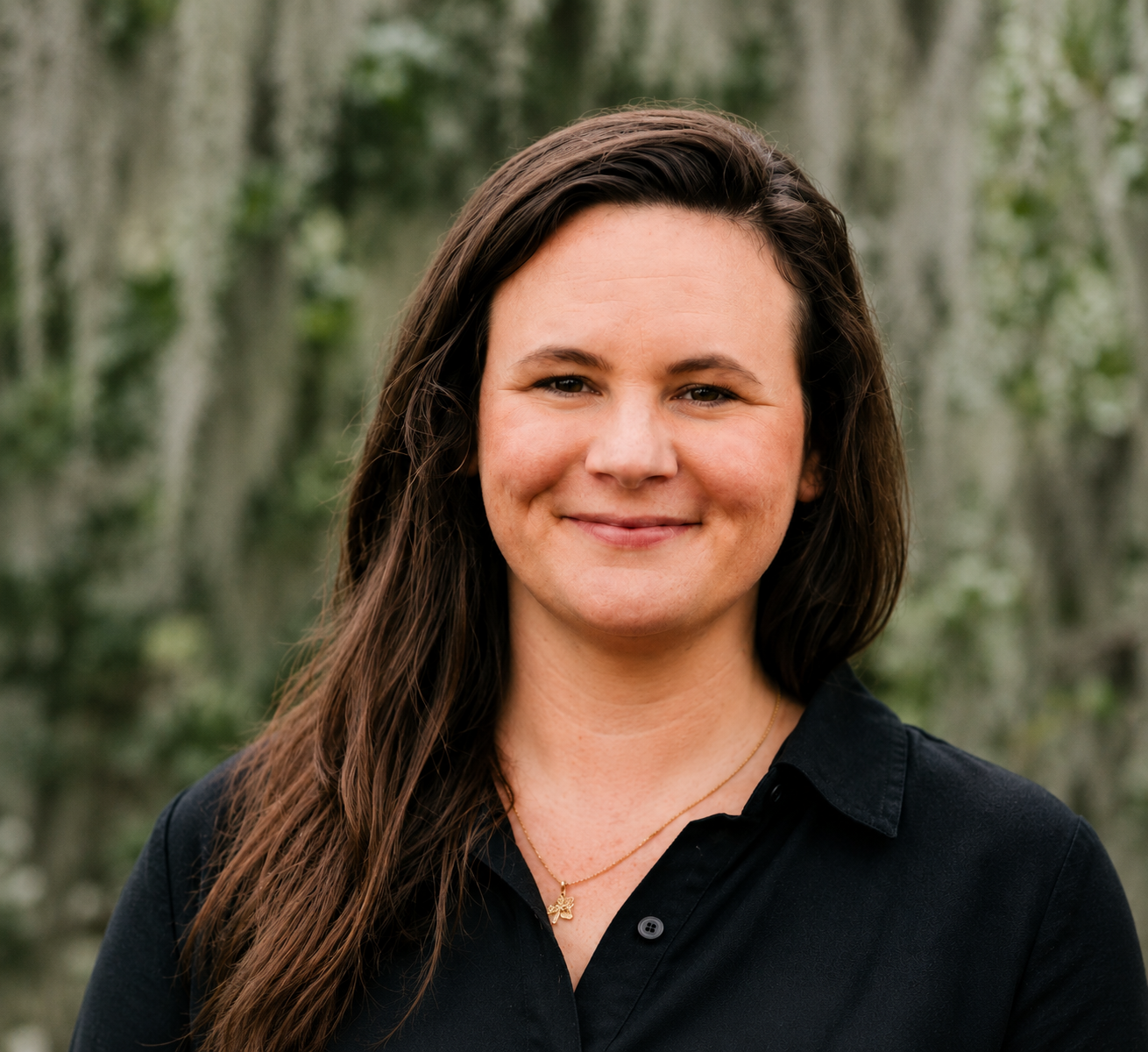A woman with long brown hair and a black shirt, smiling outdoors in front of blurred trees.