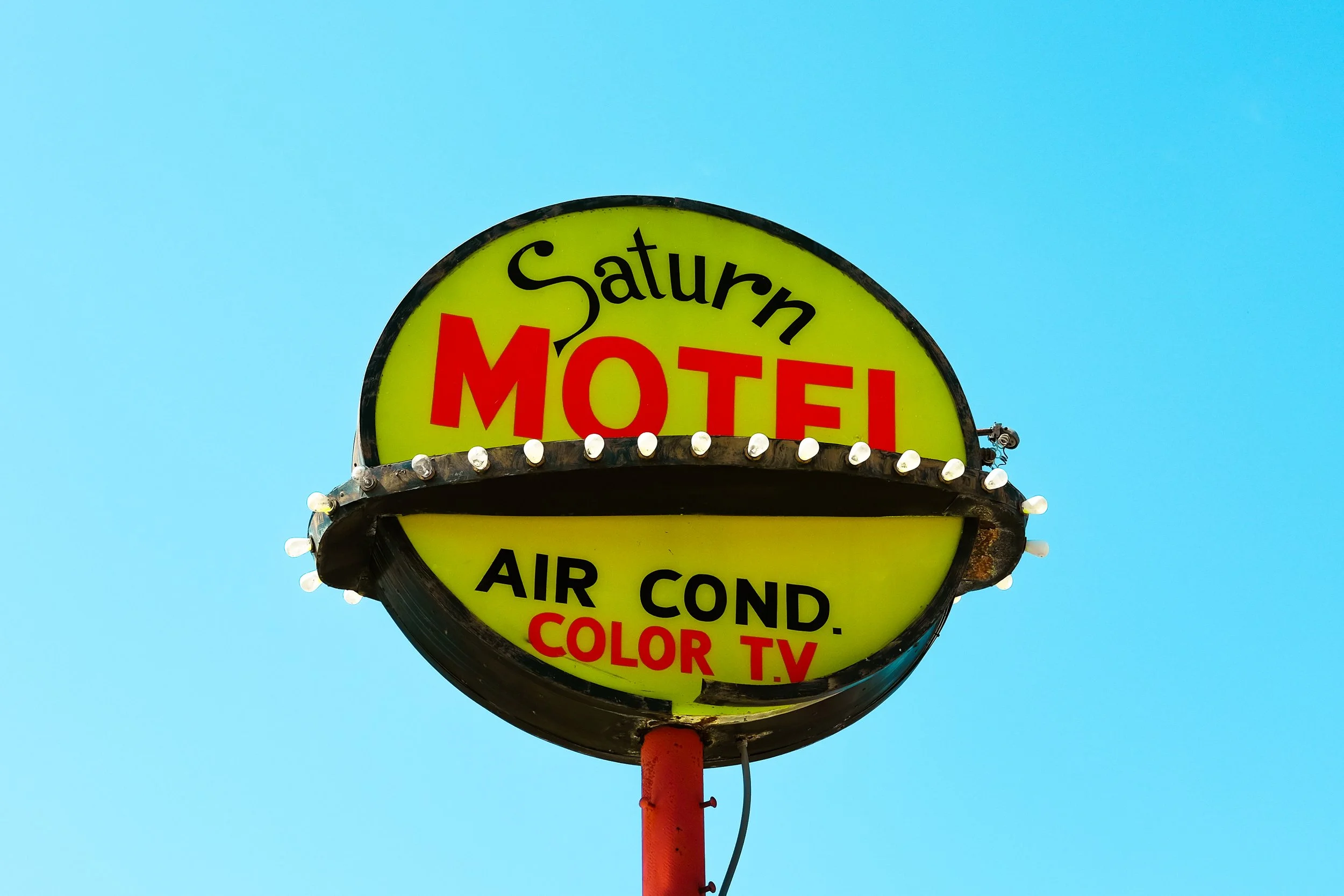 A vintage neon sign that reads 'Salturn Motel' with additional text about air conditioning, color TV, and cola, mounted on a red pole against a clear blue sky.