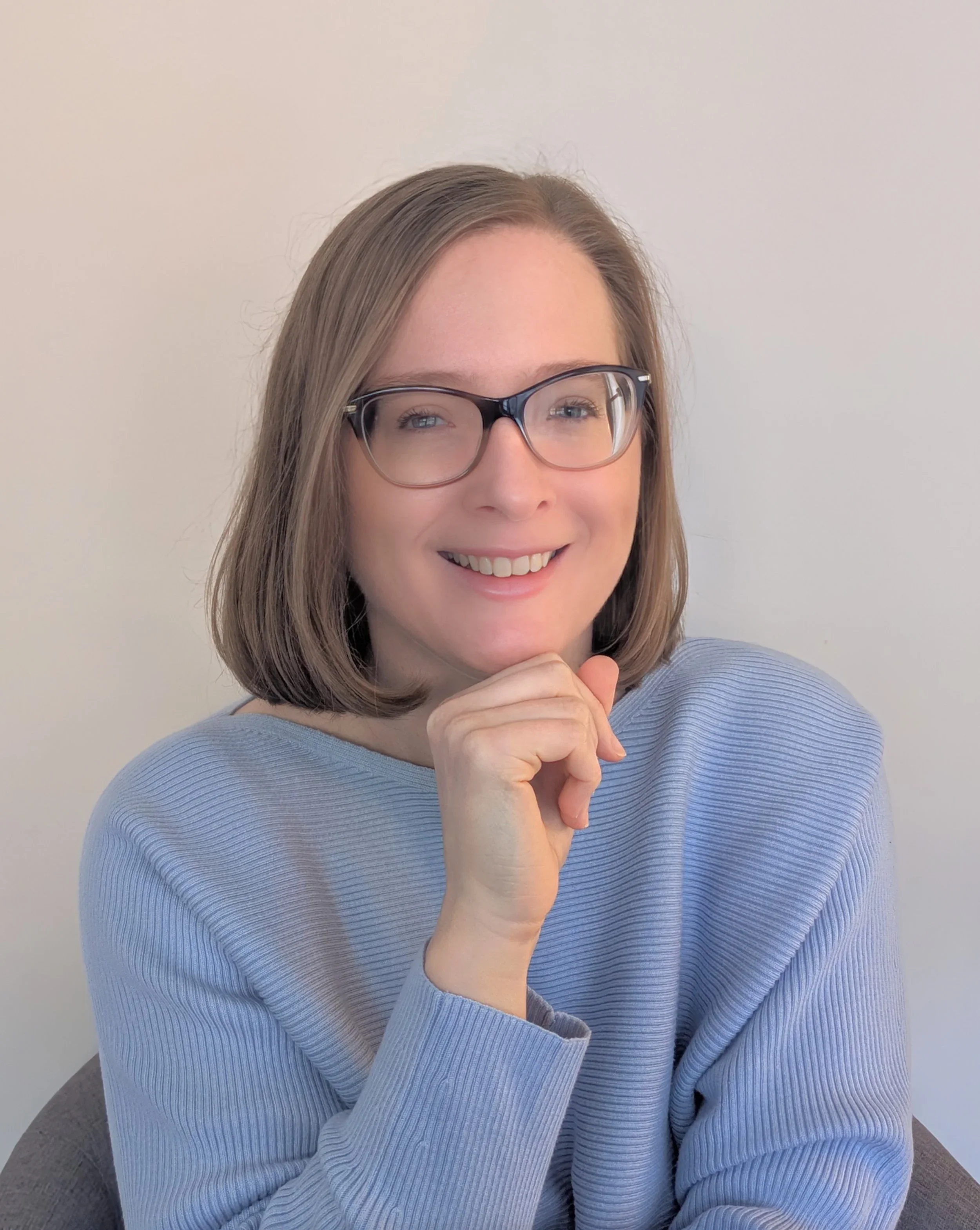 A woman with shoulder-length brown hair, glasses, and a light blue sweater, smiling and resting her chin on her hand in front of a plain white wall.
