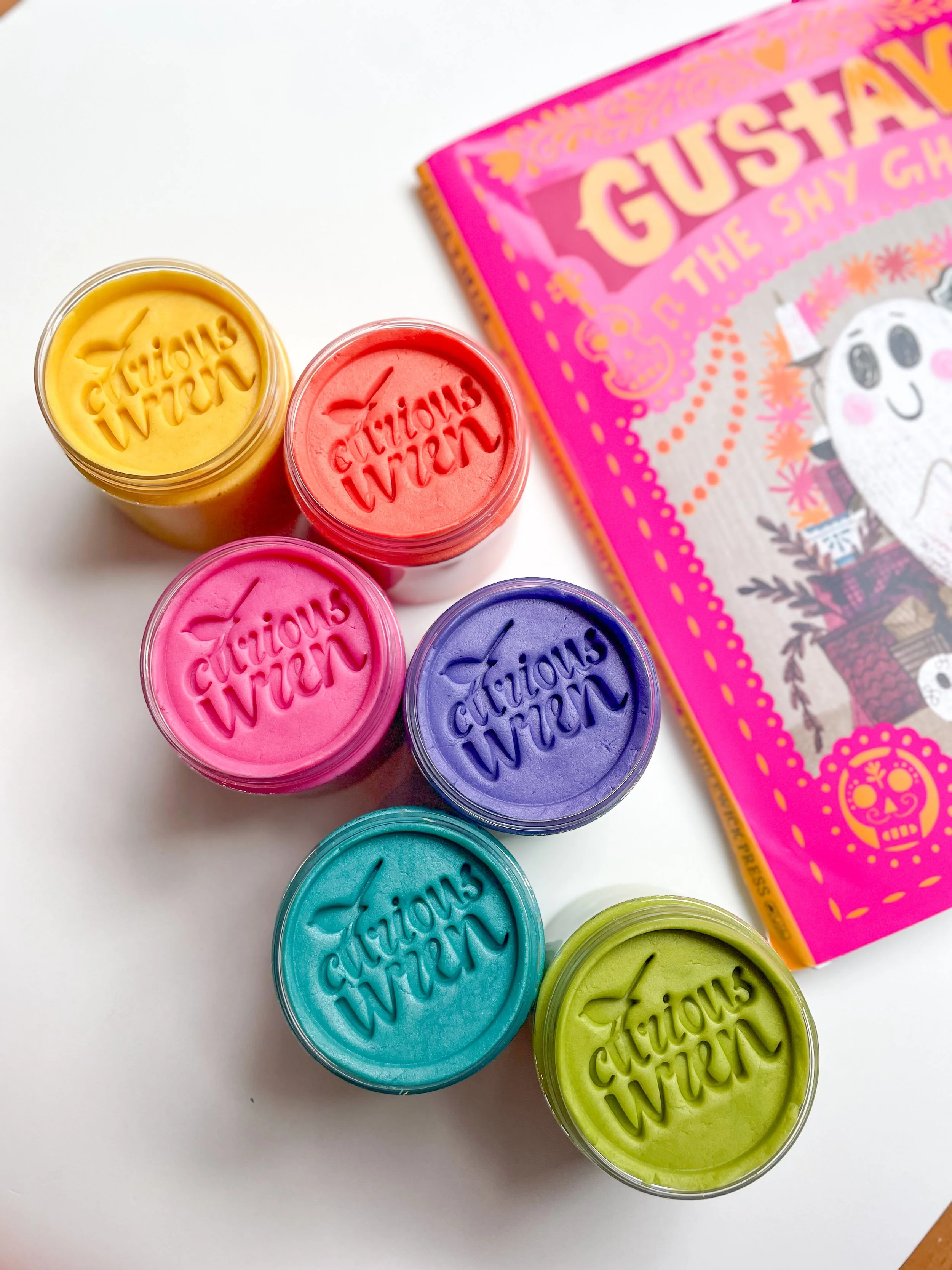 Six jars of colorful playdough arranged in a semicircle next to a brightly colored print book titled 'Gustavo the Shy Ghost' with a cartoon ghost on the cover.