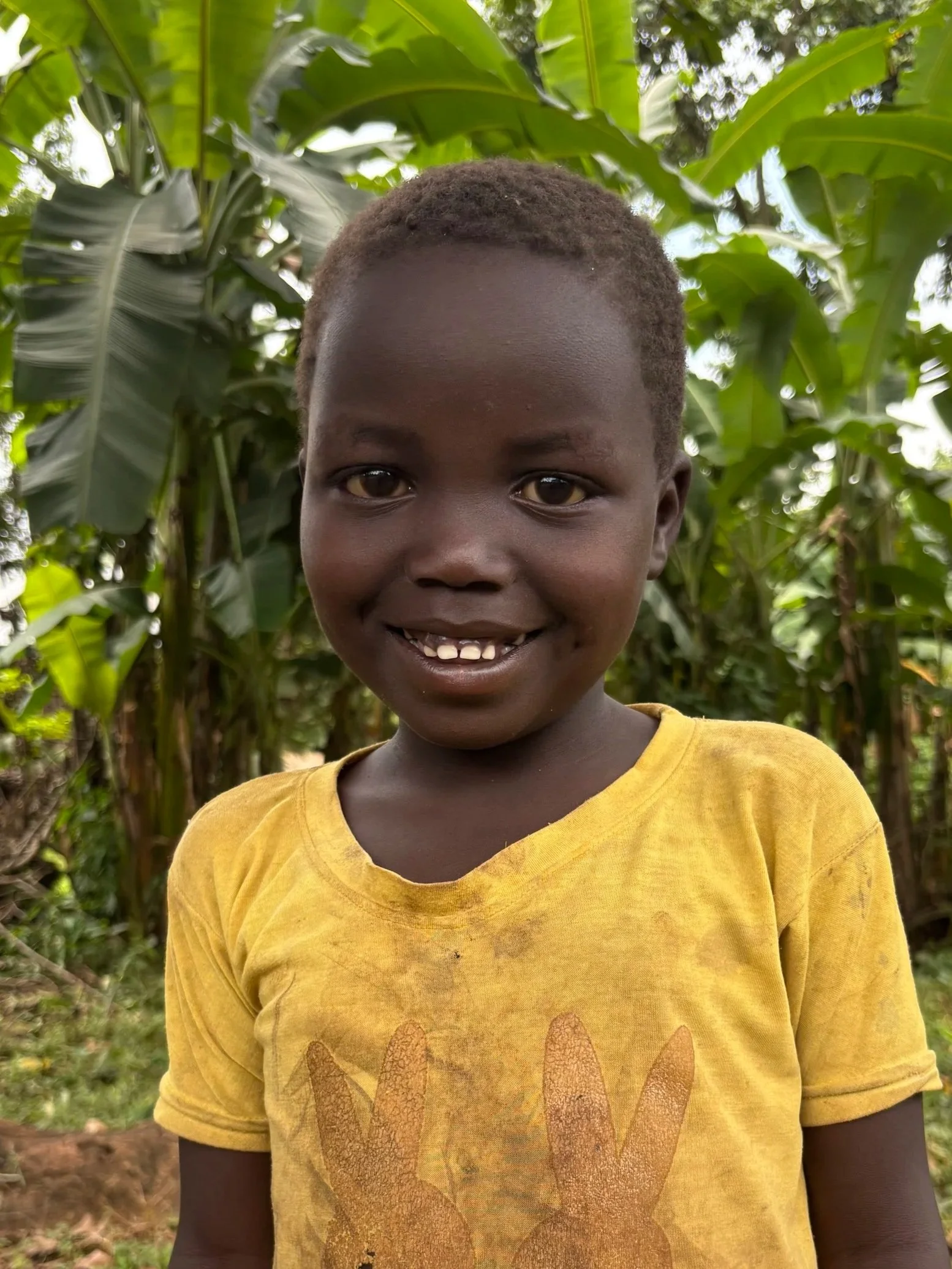 Close-up of a young boy smiling, showing teeth, wearing a yellow T-shirt with a graphic of a paw print, standing outdoors with green leafy plants in the background.