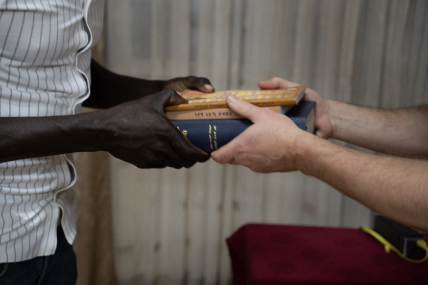 Two people exchanging a stack of books, one with dark skin and the other with light skin, against a wooden background.