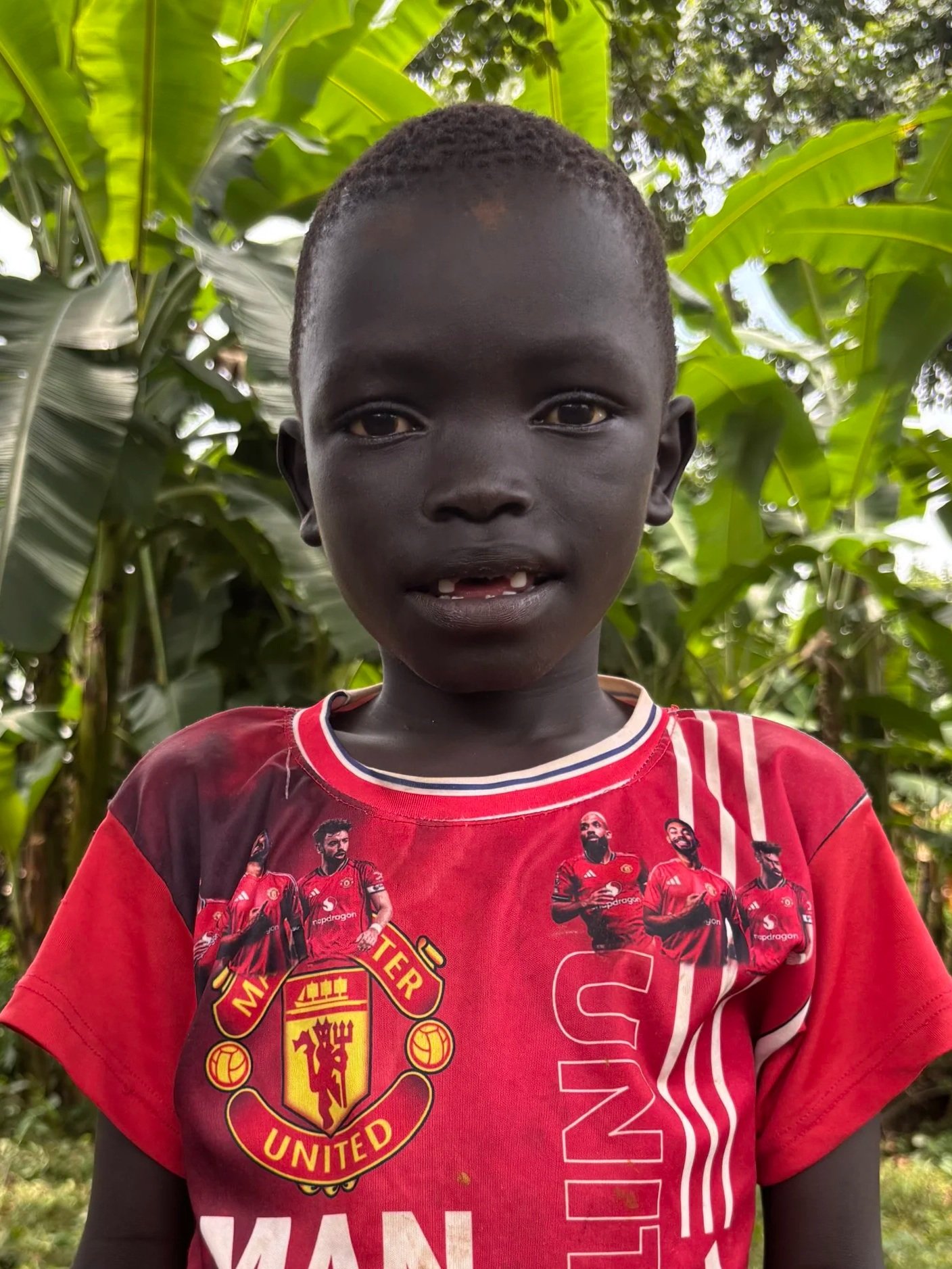 Young Black boy wearing a red Manchester United soccer jersey standing outdoors among green banana leaves.