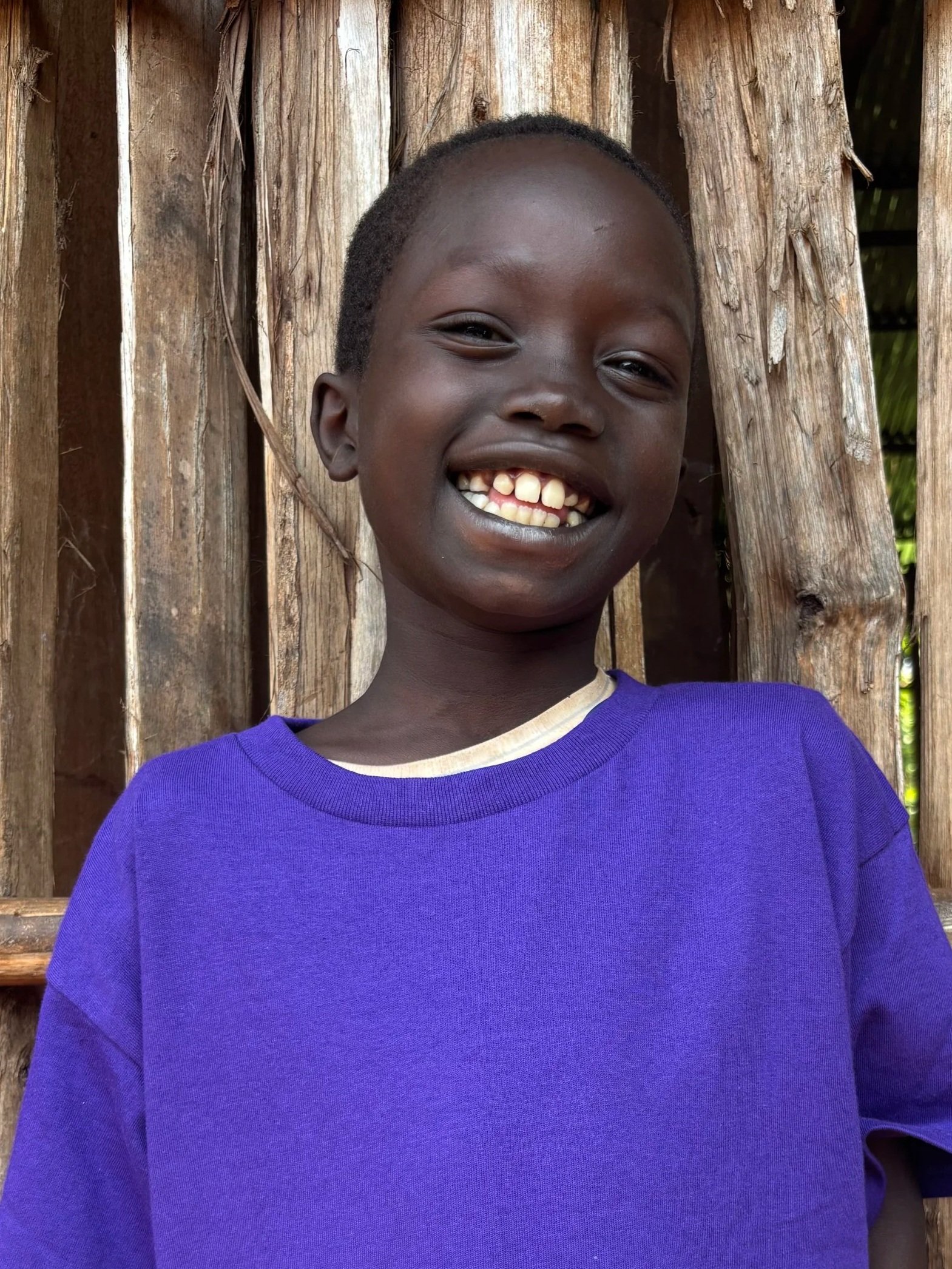 A young boy with short dark hair and a big smile showing gaps in his teeth, wearing a purple shirt, standing in front of a wooden fence.
