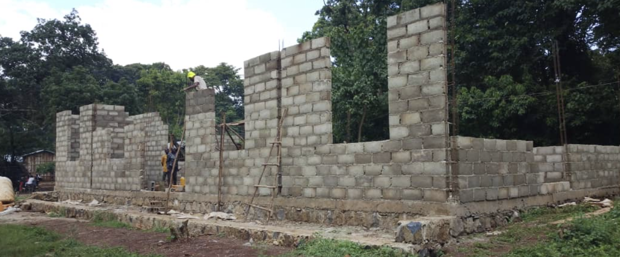 Construction site with workers building a concrete block wall, surrounded by trees, scaffolding, and construction materials.