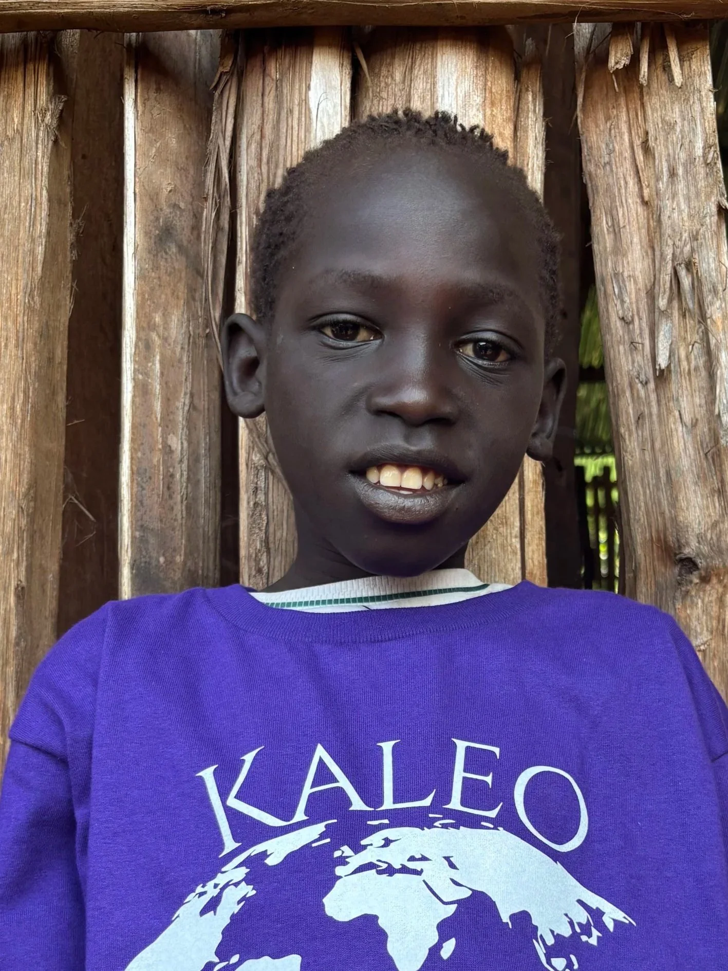 A young boy with dark skin and short curly hair, smiling slightly, standing in front of a wooden backdrop. He is wearing a purple shirt with a print of the word 'KALEO' and a globe illustration.