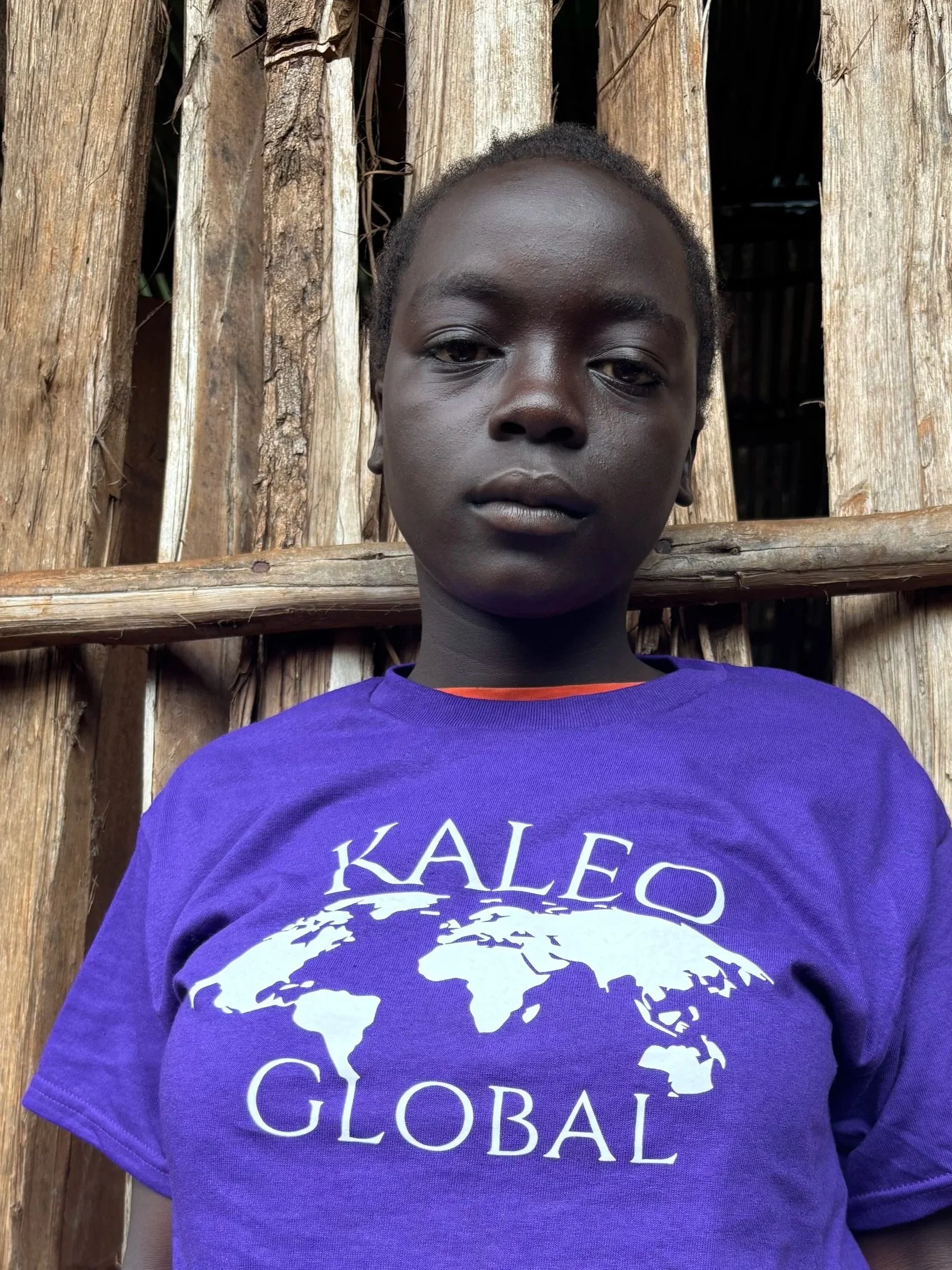 A young person in a purple T-shirt with the words 'KALEO GLOBAL' and a globe logo, standing in front of a wooden structure.