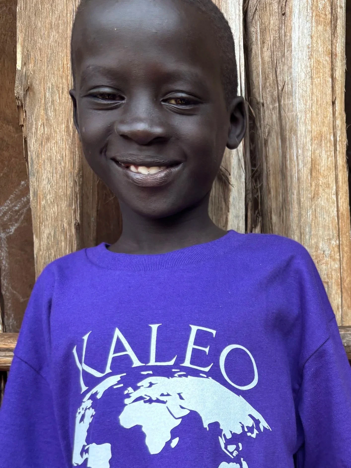 A young boy with dark skin smiling, standing in front of a wooden background, wearing a purple shirt with a white graphic of a lion and the text 'JALEO' on it.
