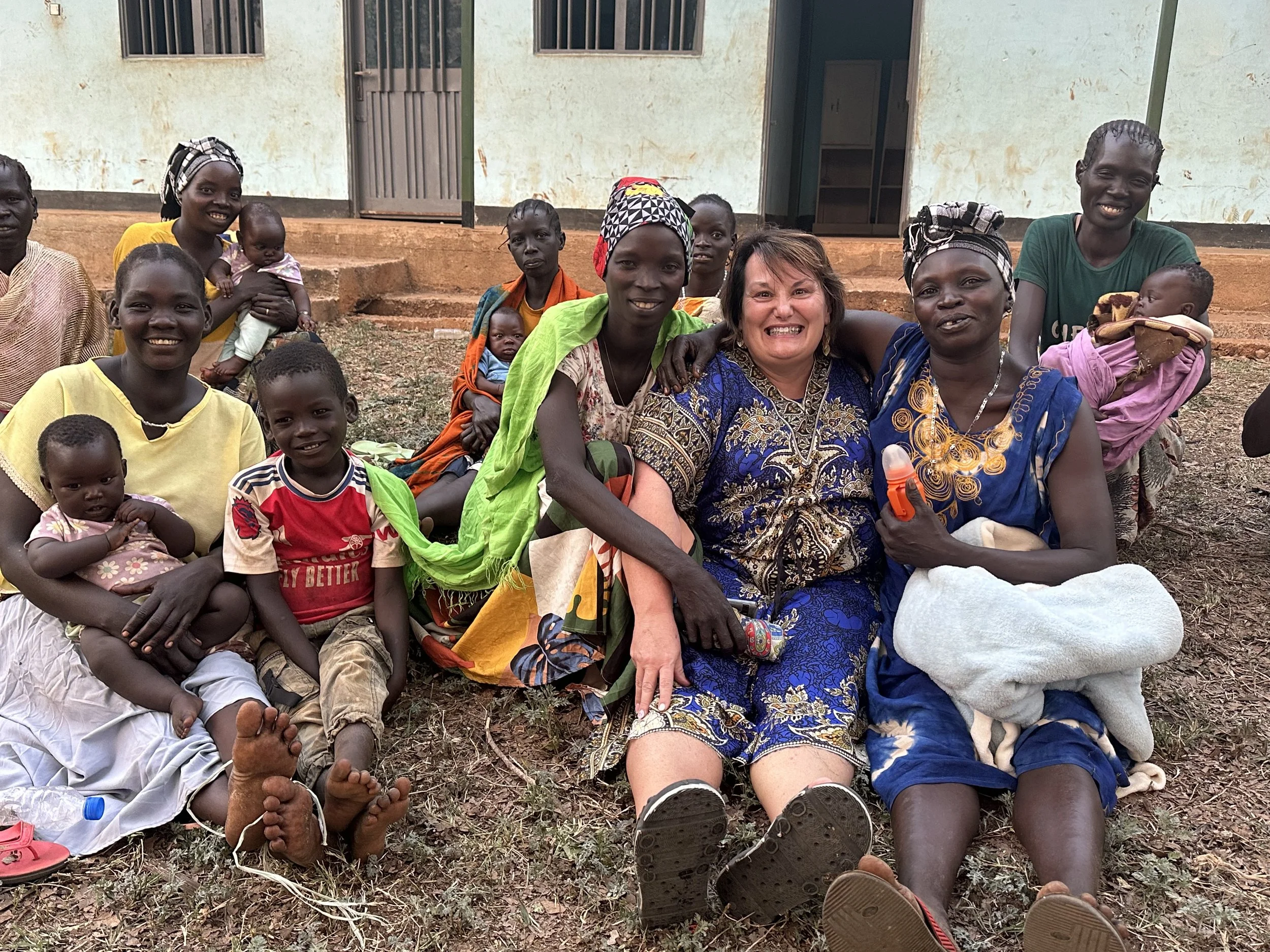 A group of women and children sitting outside on dirt ground in front of a house, smiling at the camera.