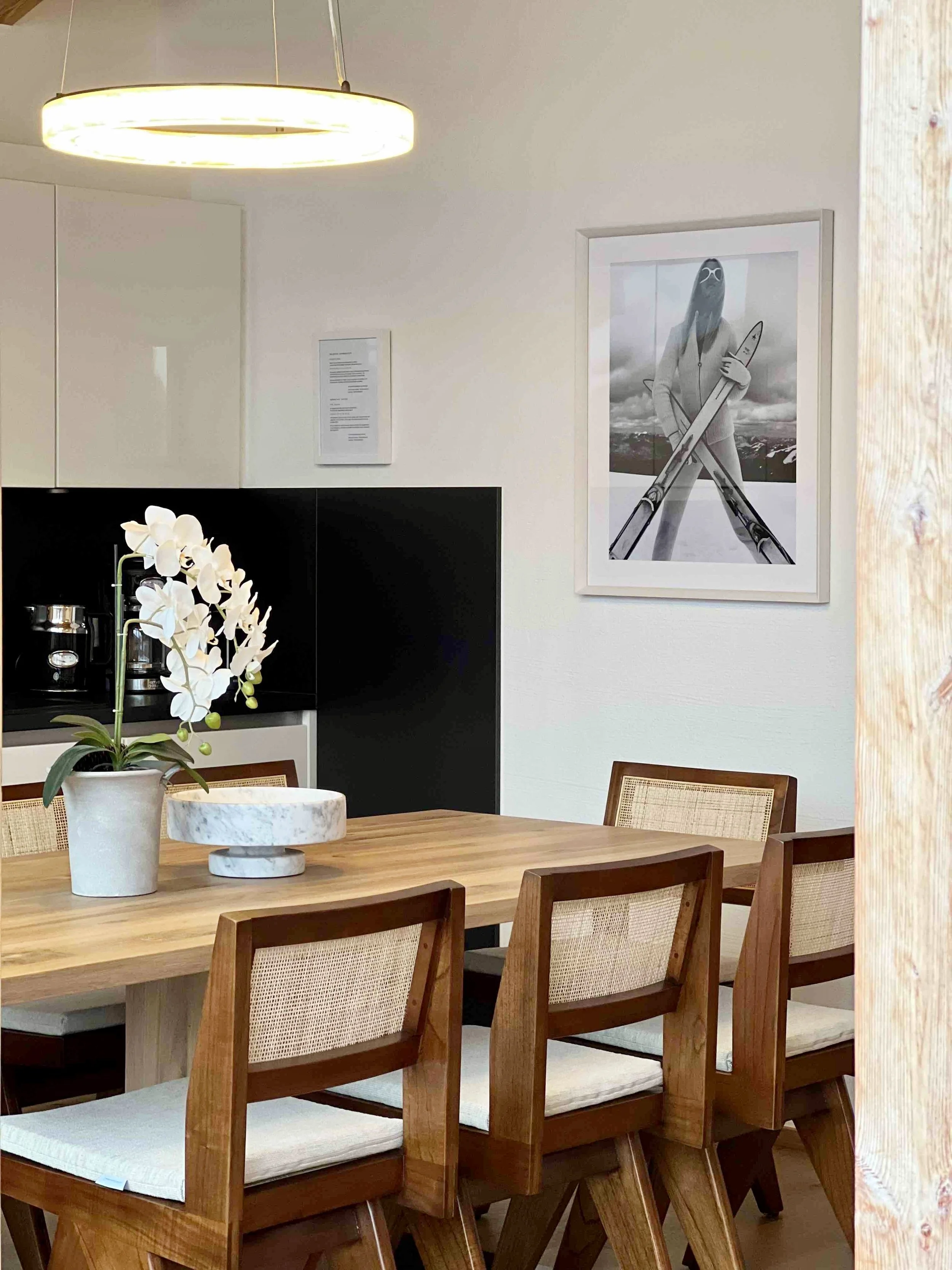 Ein Esszimmer mit einem Holztisch, Stühlen und einer weißen Orchidee in einer Vase, an der Wand hängt ein Poster mit einer animierten Person mit Skiern und Skistock in einer winterlichen Berglandschaft. Über dem Tisch hängt eine moderne Deckenlampe.