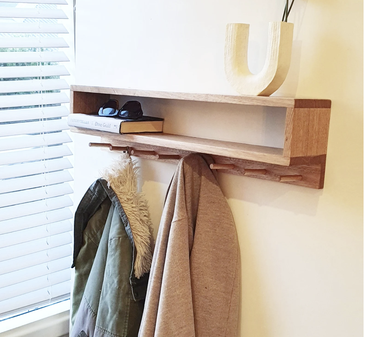 A wall-mounted wooden coat rack with hooks holding jackets, a shelf with sunglasses and books, and a decorative 'U' shaped vase on top, with a window with blinds in the background.