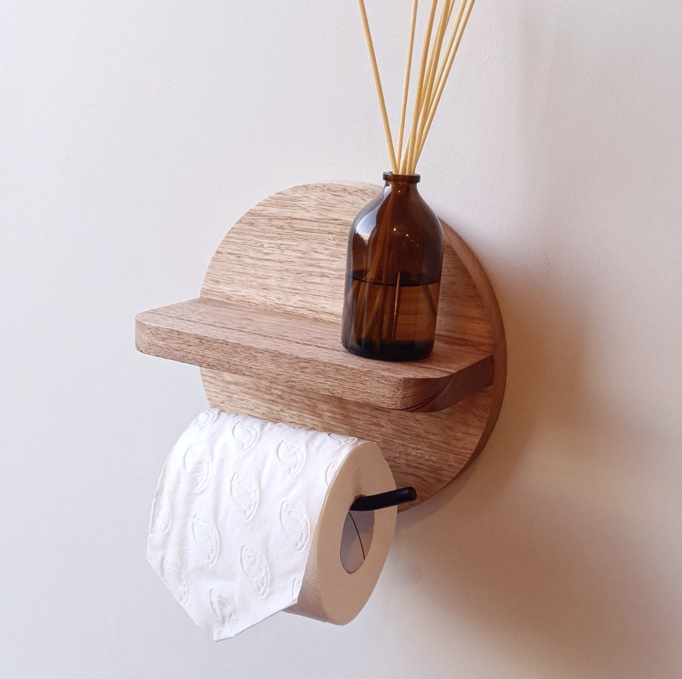 A wall-mounted wooden shelf with a small ledge, holding a brown glass vase with reed sticks, and a paper towel roll hanging from a black holder underneath.