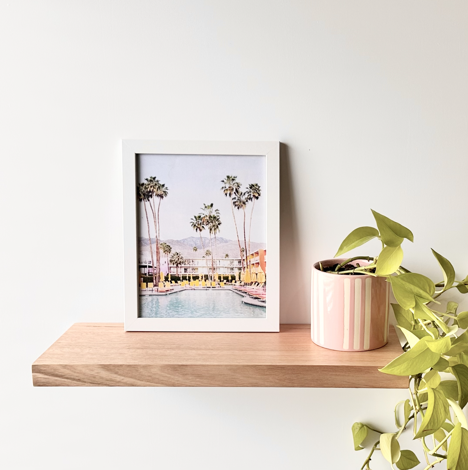 A white picture frame with a photo of palm trees and a hotel pool, sitting on a wooden shelf next to a pink striped planter with green houseplants against a white wall.