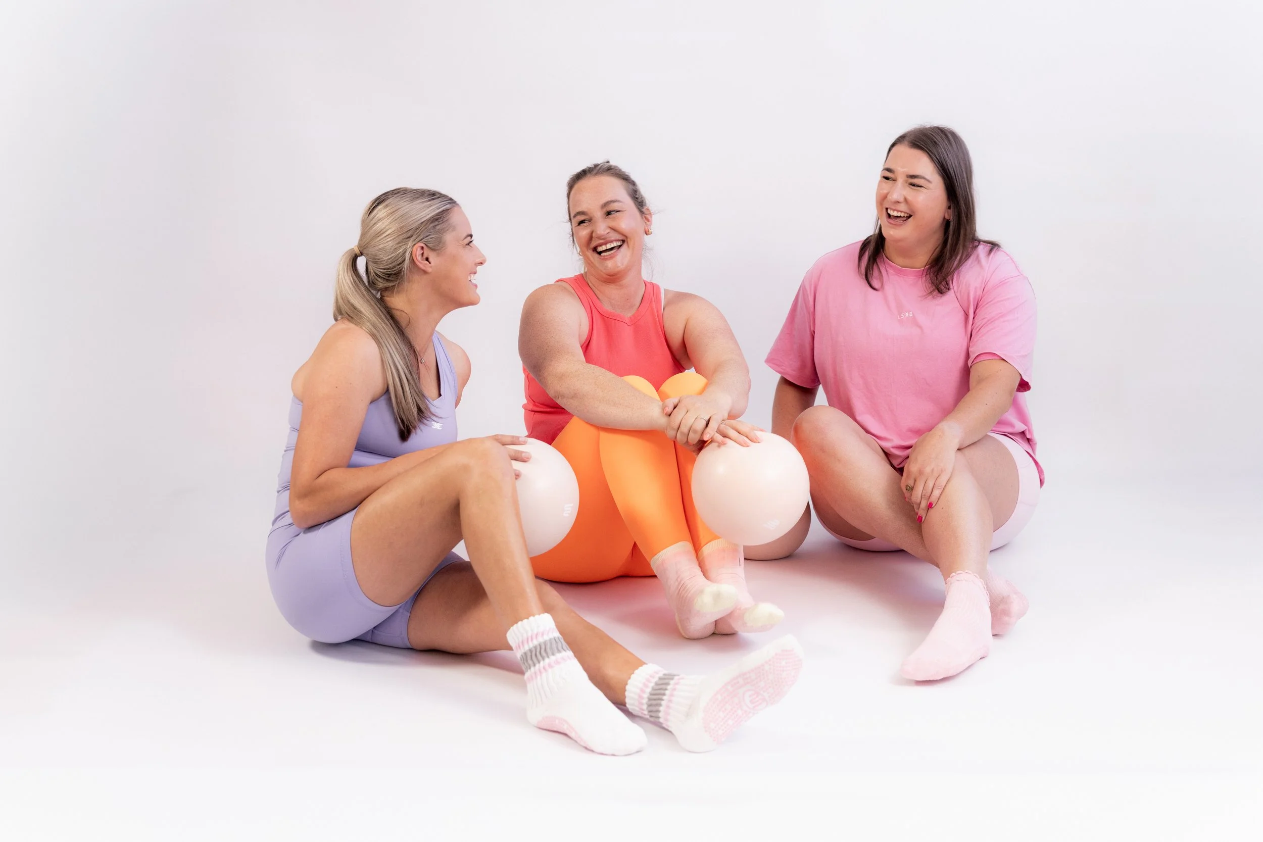  A woman in a yellow tank top is focused on exercising with a Pilates reformer, surrounded by equipment in a room with wooden floors and a shelf with colorful pillows.