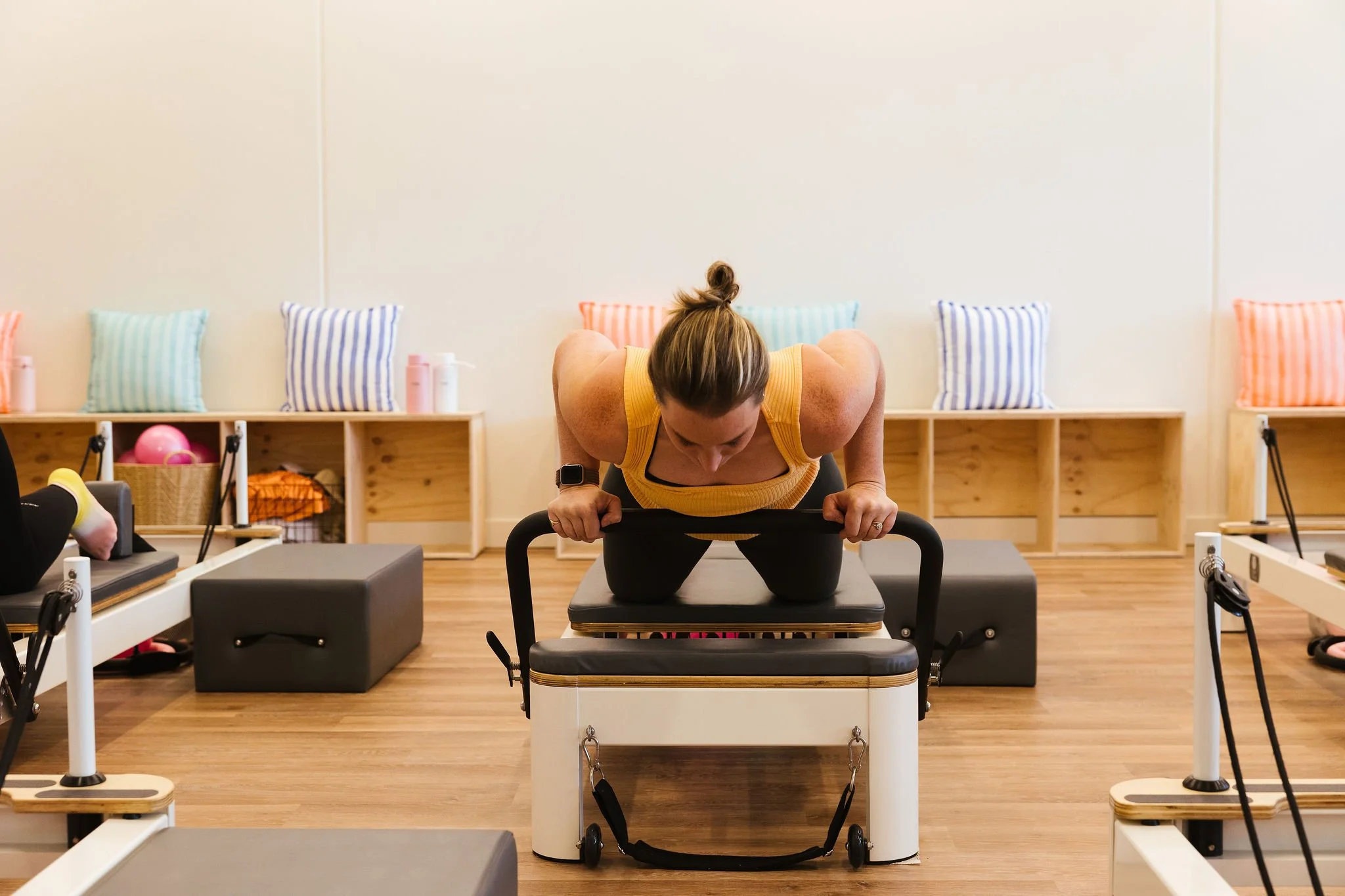 A woman in a yellow tank top is focused on exercising with a Pilates reformer, surrounded by equipment in a room with wooden floors and a shelf with colorful pillows.