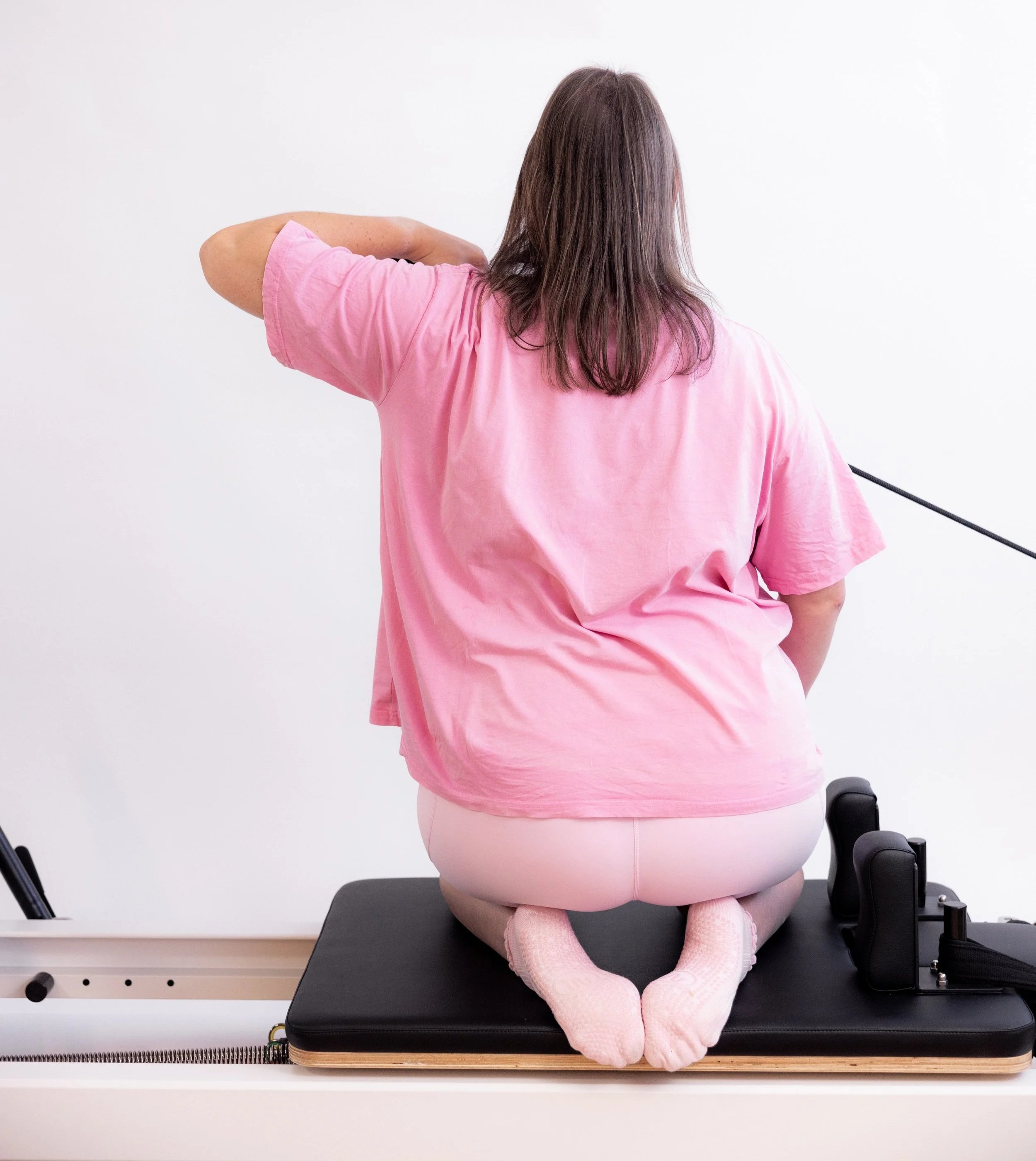  Close-up of a person holding a pink Pilates ring in one hand and a rolled-up exercise band in the other against a backdrop of grey leggings and a neutral wall.