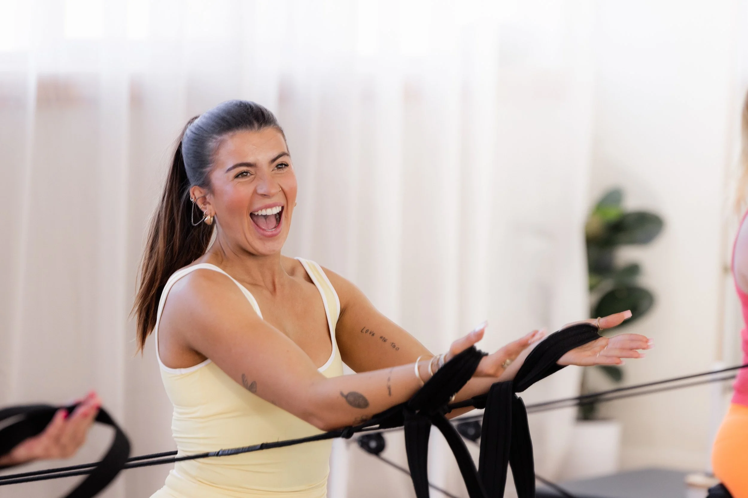Woman with dark hair in a ponytail smiling and laughing while doing reformer Pilates.