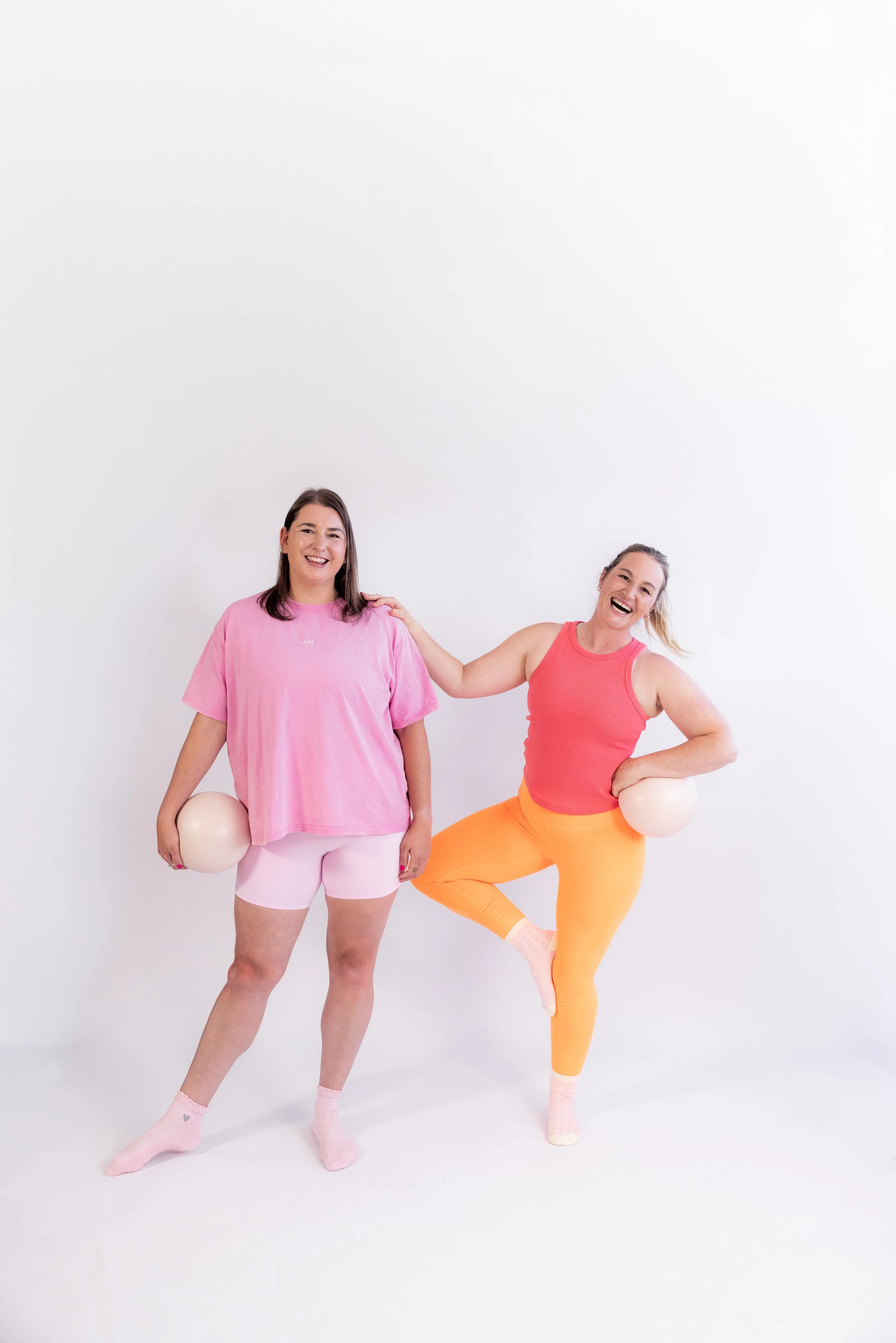 Two women in colourful workout clothes, posing with Pilates balls against a white background.