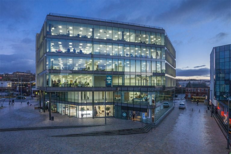 Modern office building with glass facade and multiple floors, lit up during evening time.