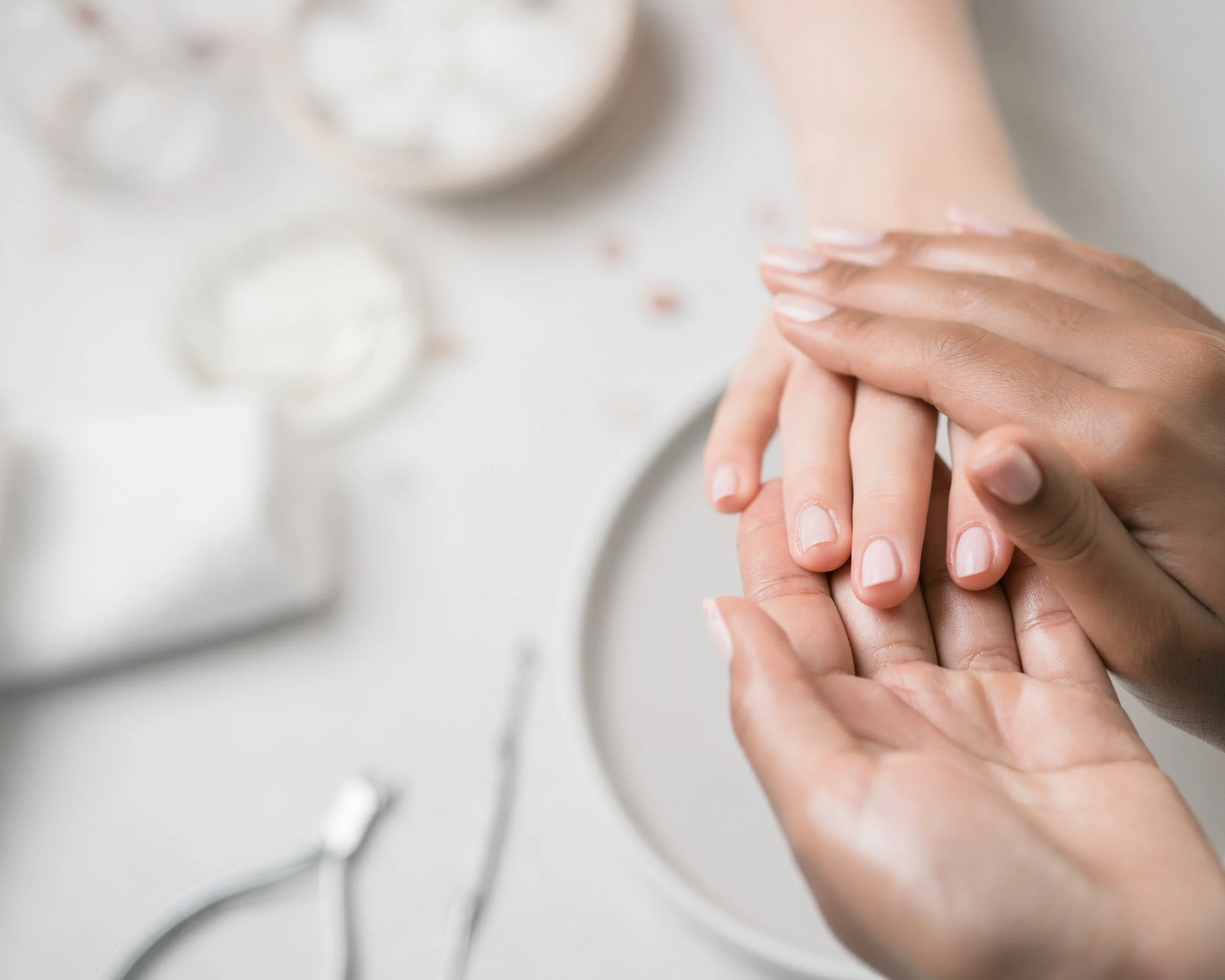 Nahaufnahme einer Handmassage, bei der eine Hand die andere sanft hält und massiert, im Hintergrund unscharfe Maniküre-Werkzeuge auf weißem Untergrund