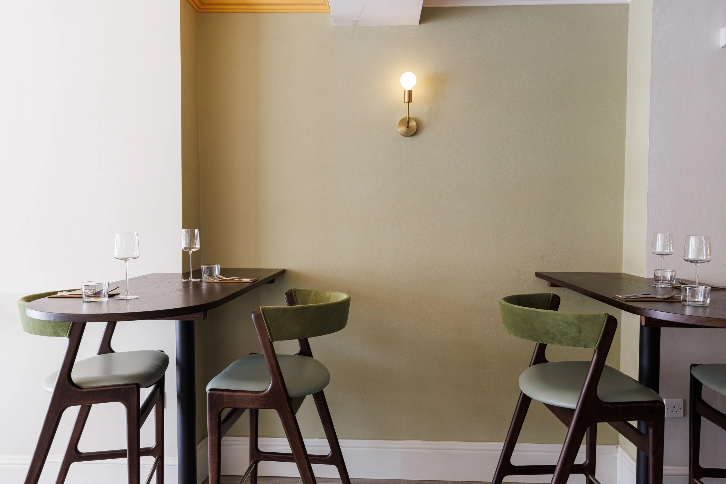 Interior of a restaurant with two dark wooden high tables, green cushioned chairs, wine glasses, and water glasses set for dining, with a wall-mounted light fixture on a beige wall.