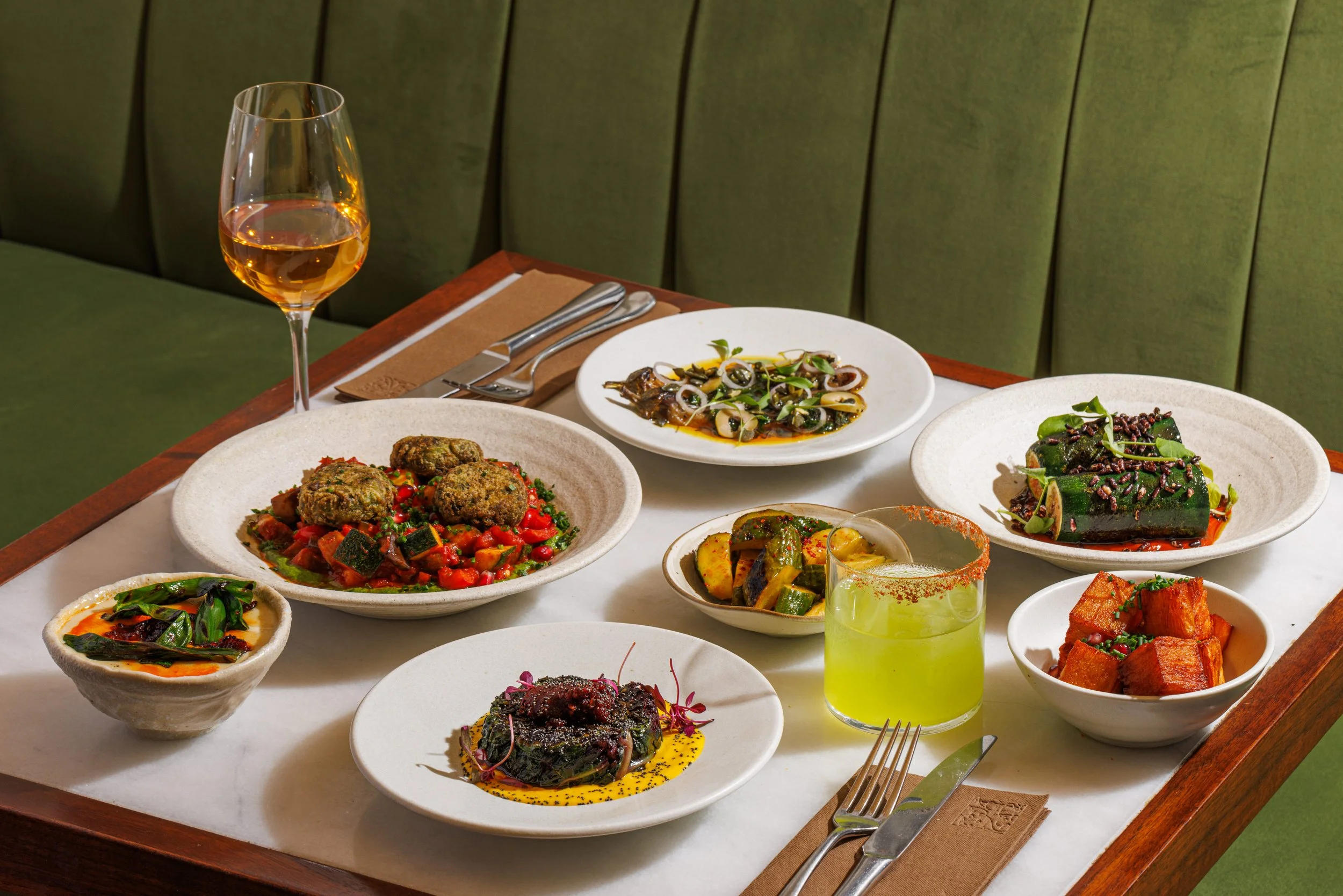 A table set with various dishes including meatballs with vegetables, grilled zucchini, stuffed zucchini rolls, roasted eggplant, and crispy tofu, along with a glass of rosé wine and a bright yellow cocktail.
