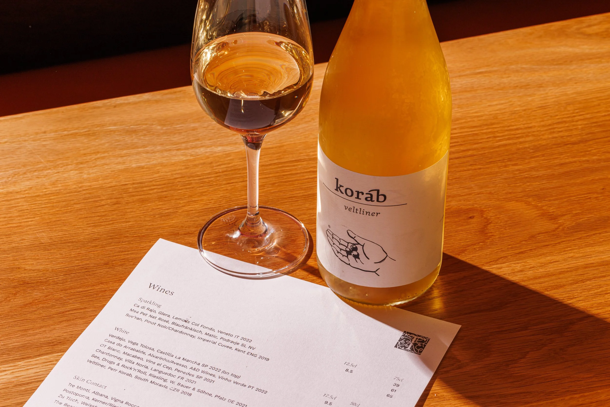 A glass of white wine and a bottle labeled "korab veltliner" on a wooden table, with a menu underneath.