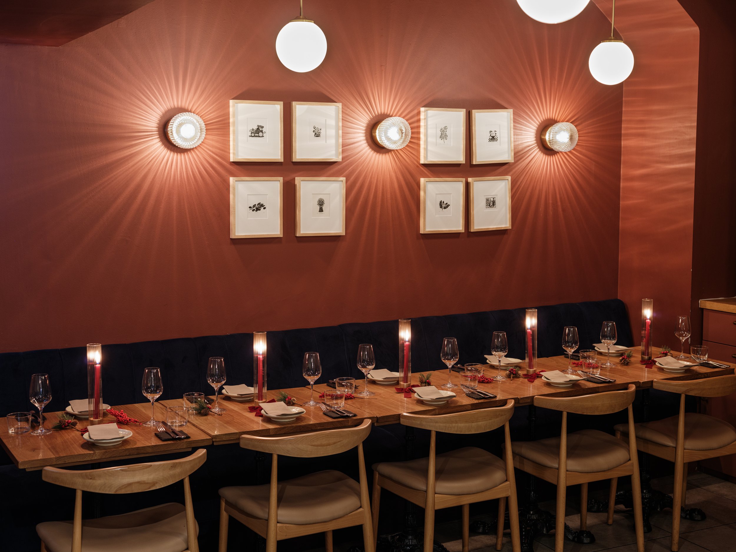 A cozy dining area with a long wooden table set with wine glasses, white plates, napkins, and decorative candles. The wall behind features eight framed black-and-white artworks arranged in two rows, and ambient lighting creates starburst patterns on 