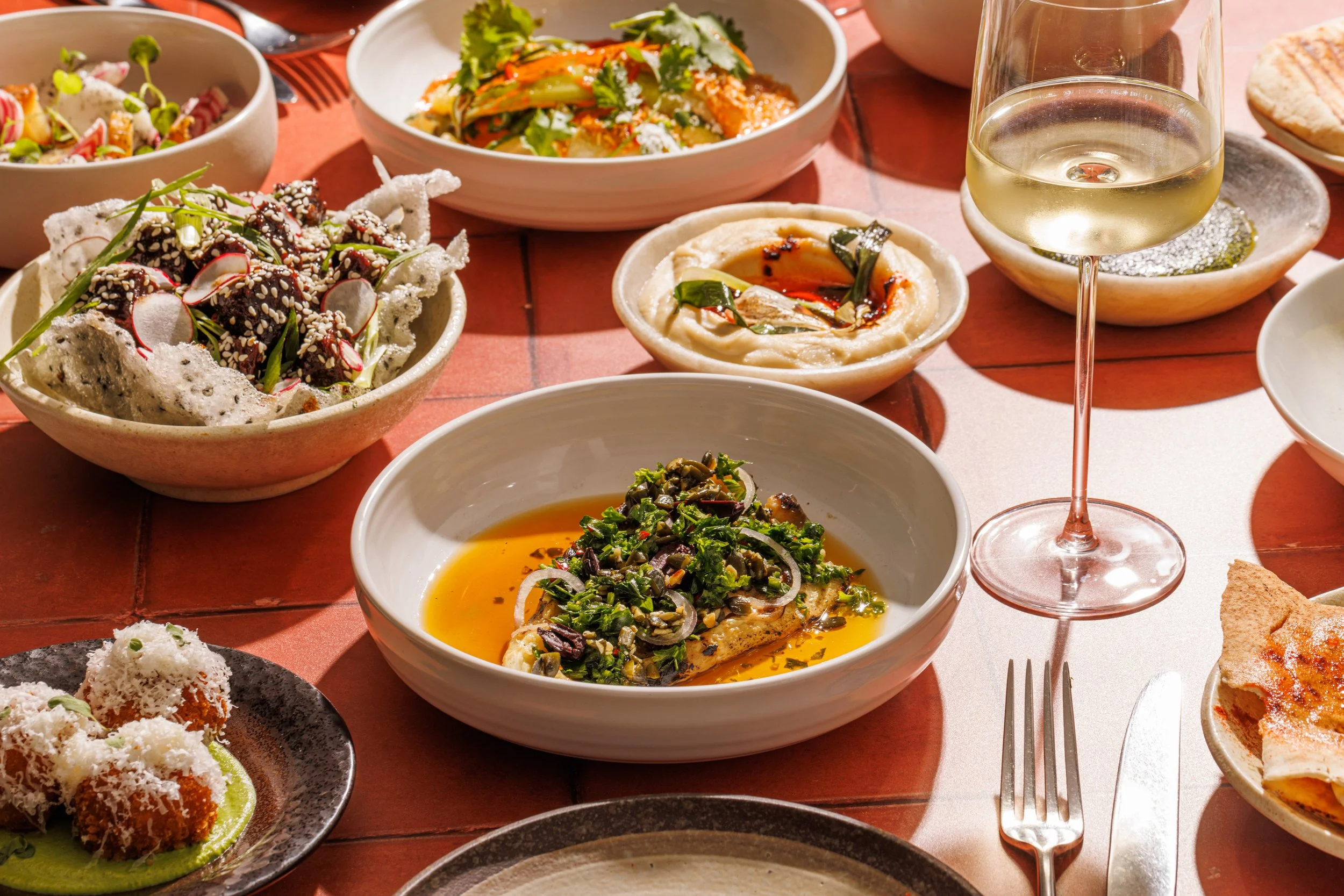 Assorted dishes on a red table, including a bowl of fish topped with herbs, a large salad, hummus with paprika and herbs, a glass of white wine, and various appetizers and side dishes.