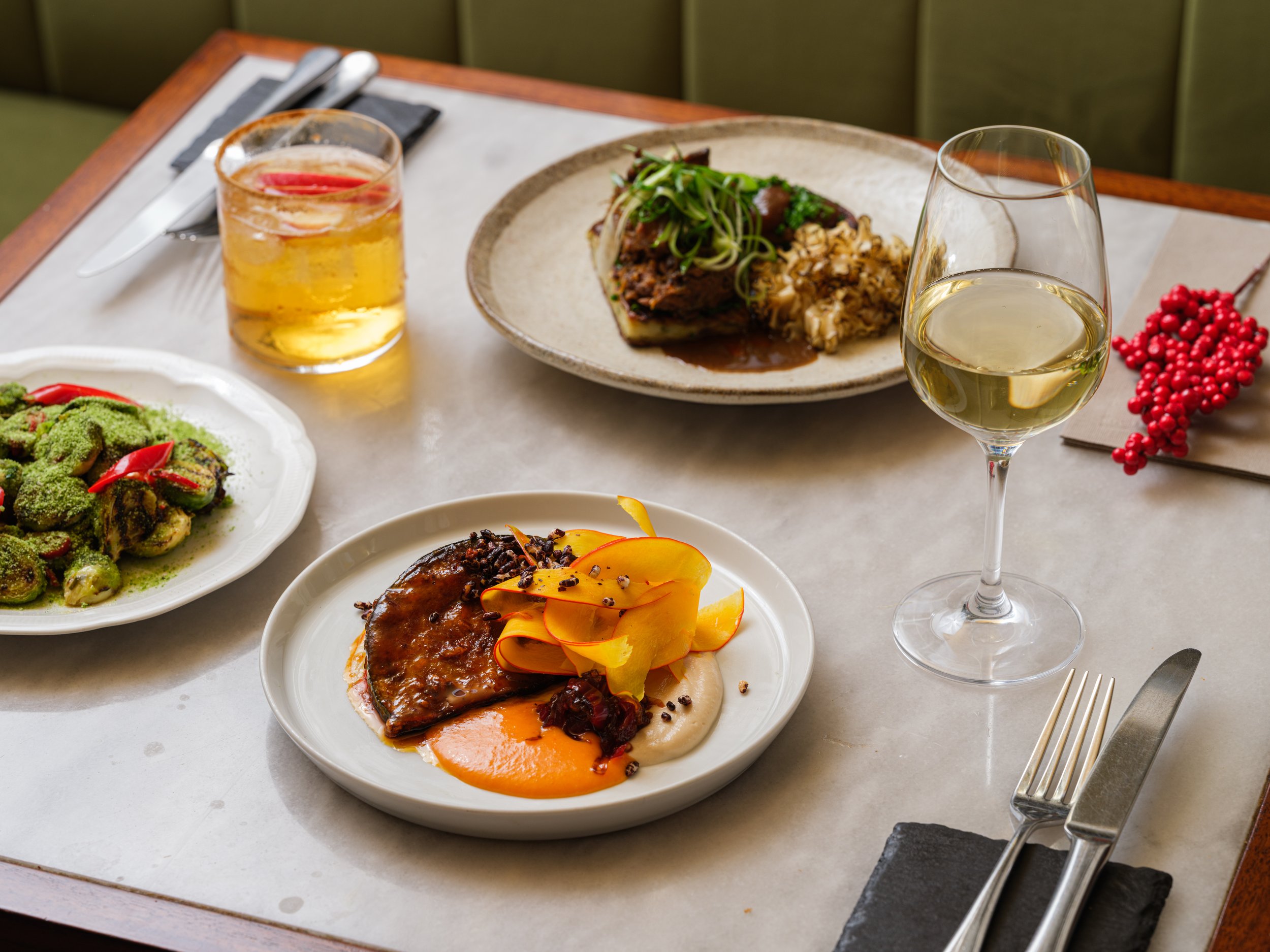 A table set with various dishes including a glass of white wine, a glass of iced beverage, a plate of vegetables, and a main course with meat and vegetables, on a white marble surface with cutlery and black napkins.