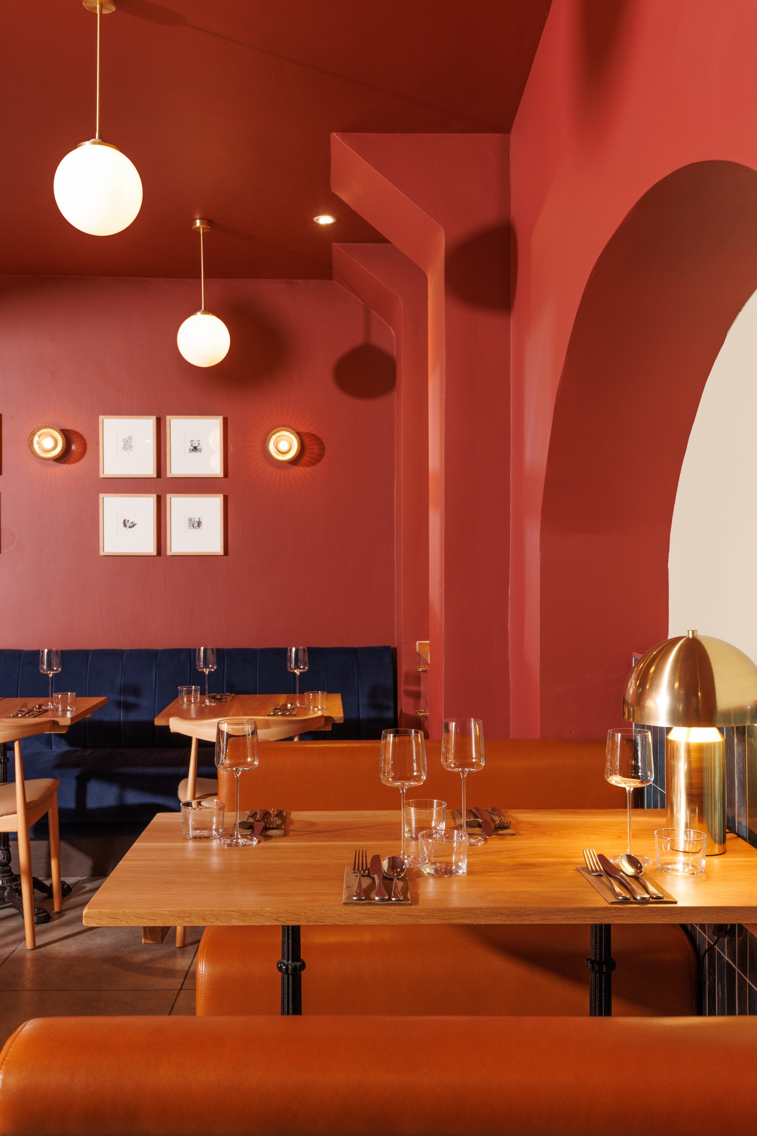 Interior of a restaurant with pink walls, pendant and wall lights, framed artwork, and tables set with glassware and cutlery.