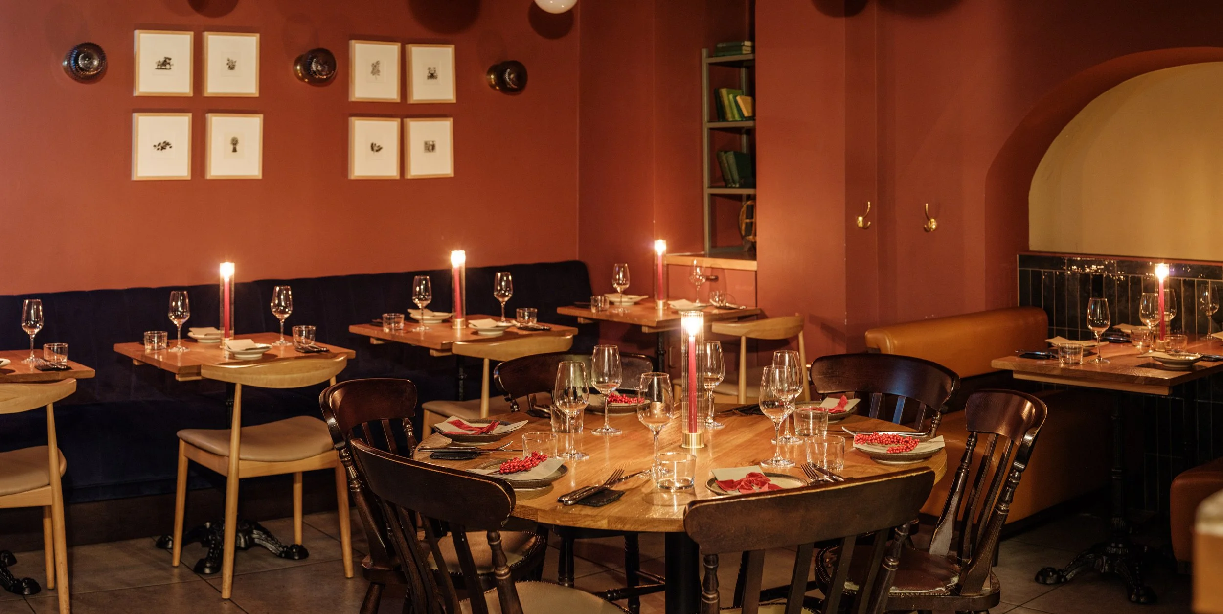 A cozy restaurant interior with wooden tables set with wine glasses, water glasses, plates, forks, knives, napkins, red candles, and red beads, pink walls, framed artwork, and bookshelves.