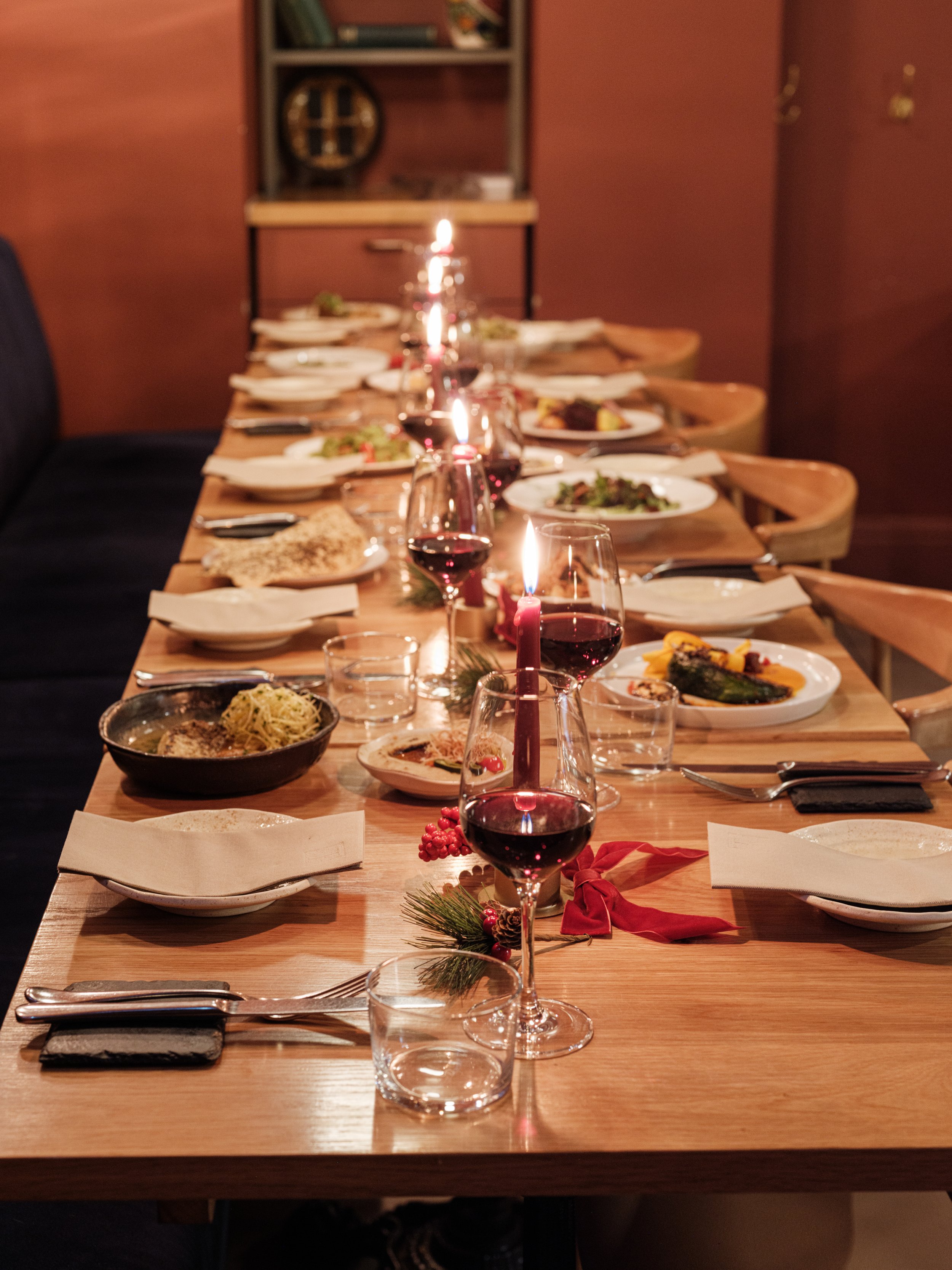 A long wooden dining table set with plates of food, glasses of red wine, and lit candles in a dimly lit room with wooden walls.
