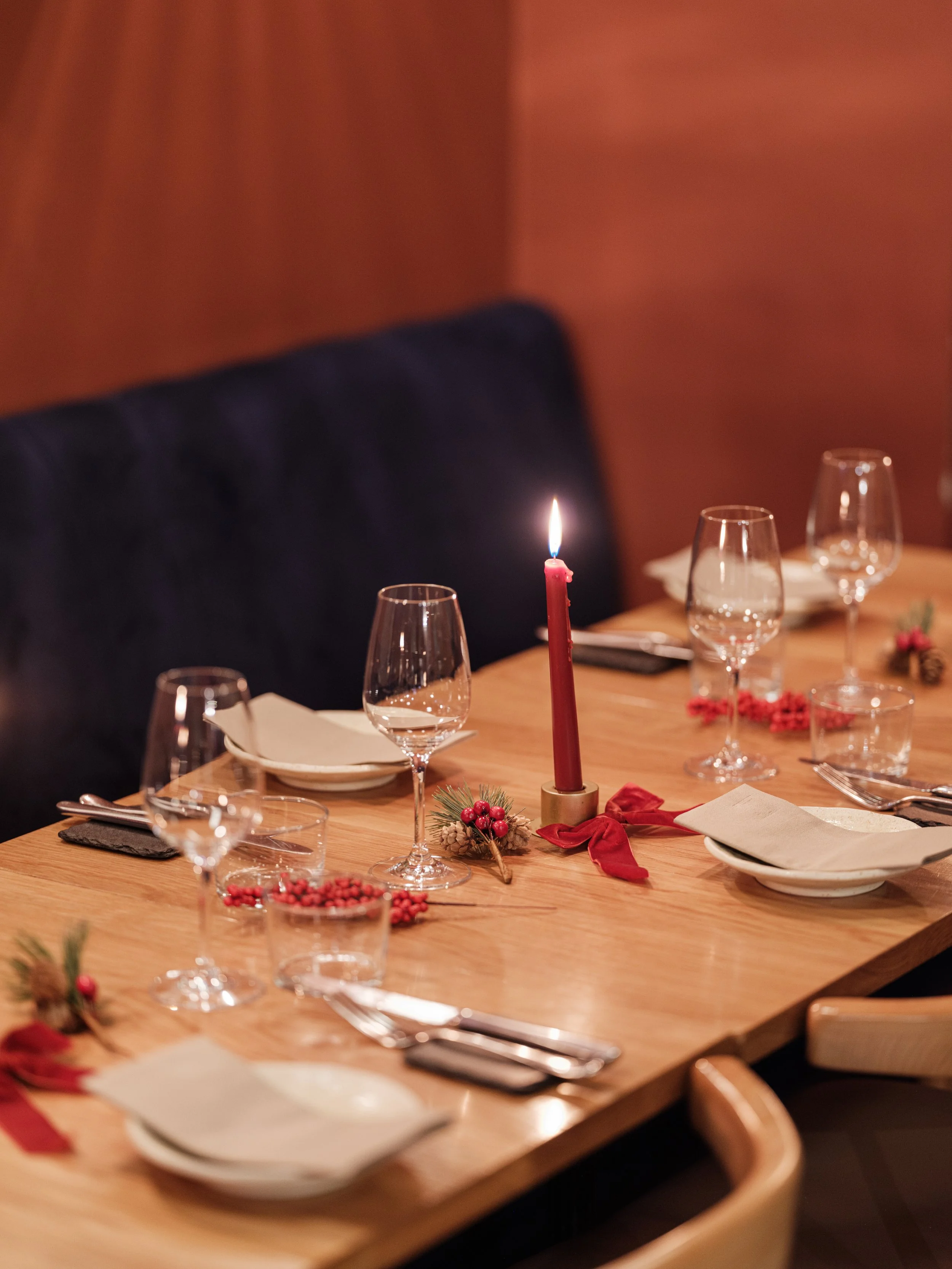 A decorated dining table with a red candle in a brass holder, surrounded by empty wine glasses, plates, silverware, and holiday-themed decorations including pinecones, artificial berries, and a red ribbon.