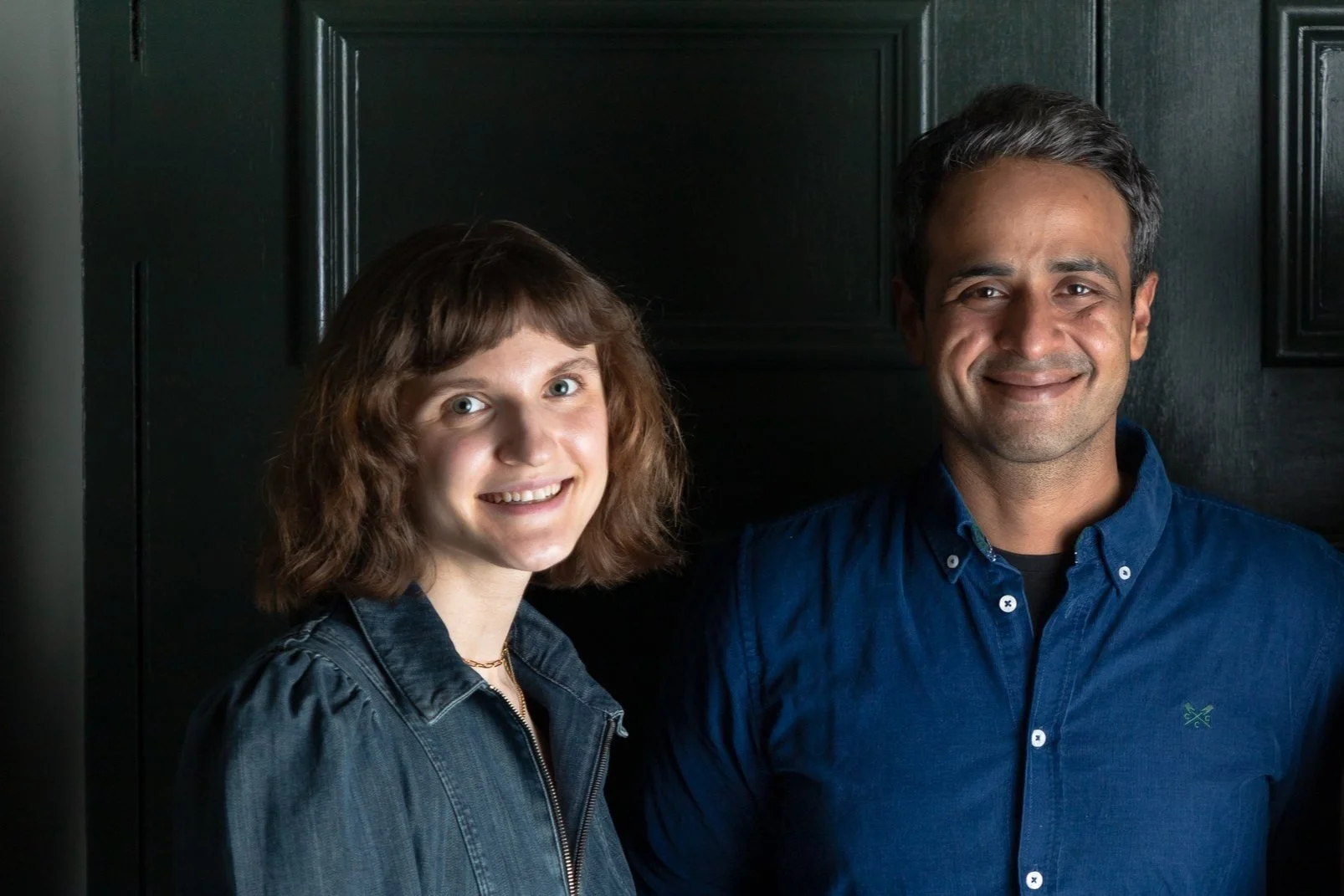 A young woman and a man standing side by side, smiling in front of a dark wood-paneled background.