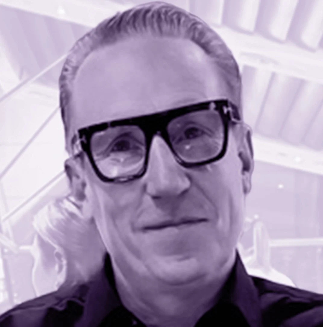 A man with combed-back hair and black-rimmed glasses, smiling, in an indoor setting with a high ceiling.