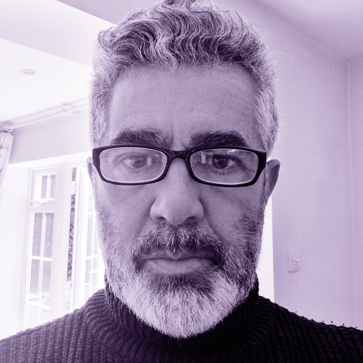 Close-up photo of a middle-aged man with gray hair, beard, and glasses, indoors with windows and curtains in the background.