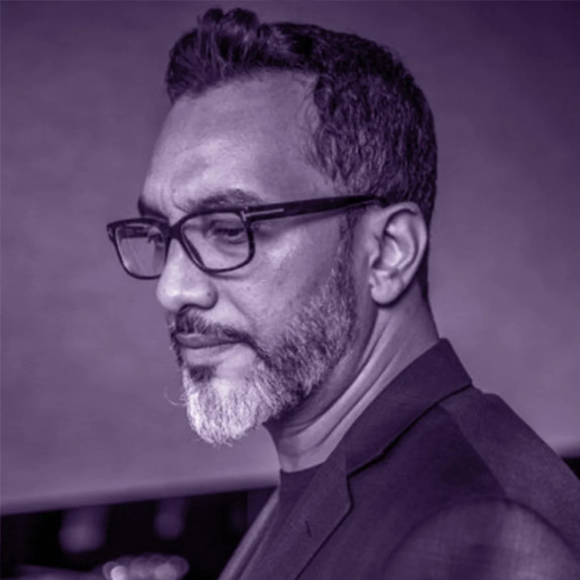 A profile portrait of a middle-aged man with glasses, short curly hair, and a beard, wearing a dark suit and a turtleneck, against a blurred background.