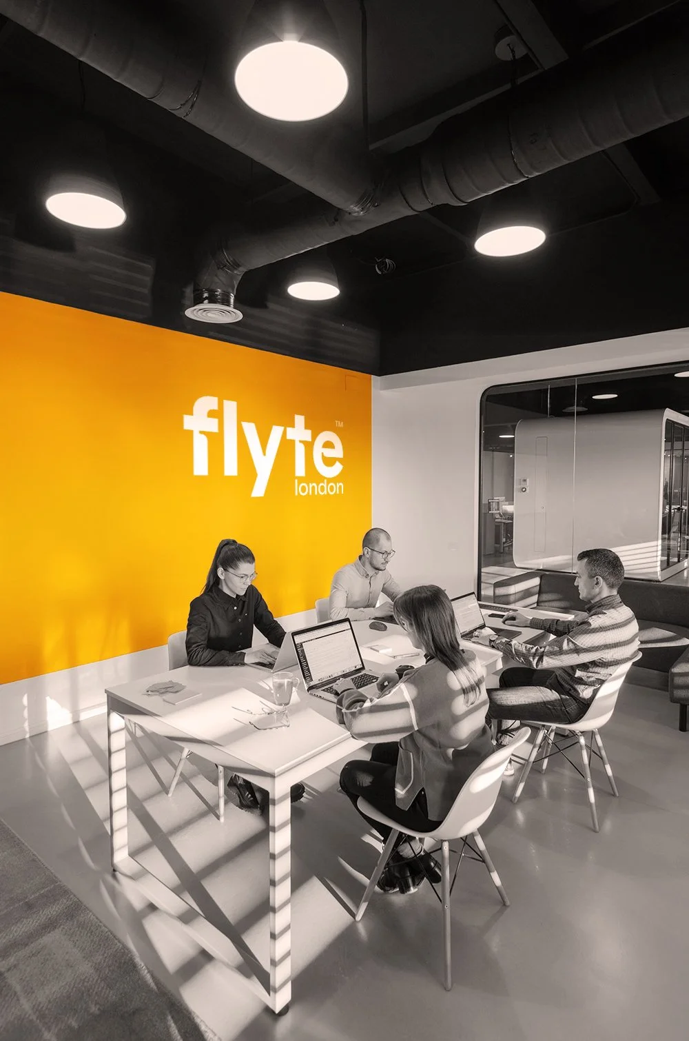 Four people sat sharing a desk in modern office location. FlyteLondon logo printed large in white against an orange wall.