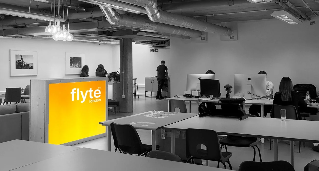 An office space with desks, computers, and people working. A large yellow sign with white text reads 'flyte london' in the foreground, and the rest of the image is mostly in black and white.