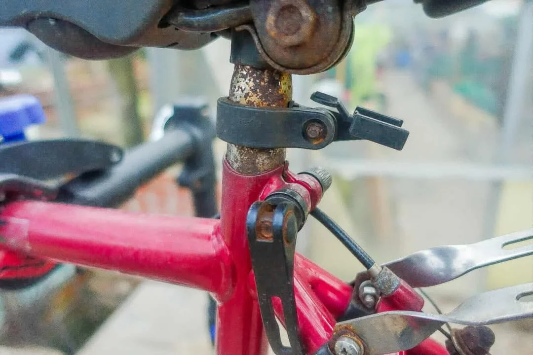 Close-up of a bicycle seat tube with a red frame, showing a rusty seat post clamp, a black quick-release seat post collar, and a black mounting bracket. The bike is rusty, with some corrosion on metal parts, and the background is blurred with outdoor scenery.