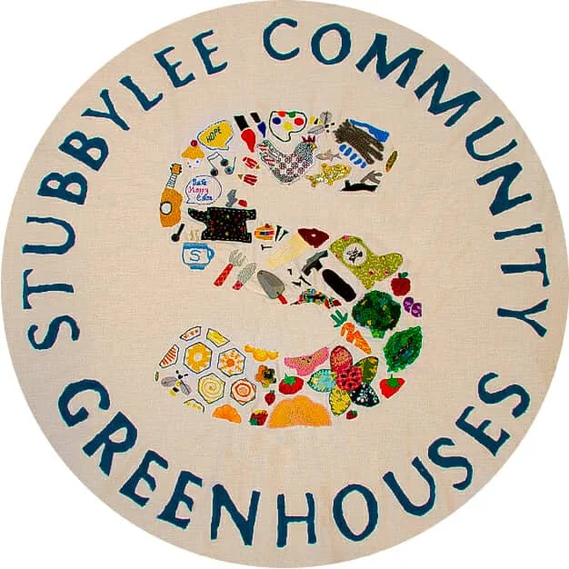 A circular fabric sign with the words 'Stubbleyee Community Greenhouses' around the edge. The center features a colorful mosaic of various objects and symbols, including flowers, vegetables, and gardening tools.