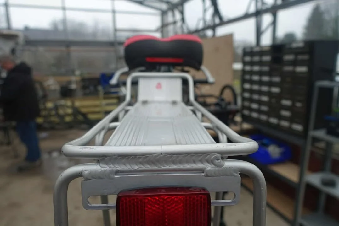 Close-up of the rear of a bicycle carrier rack with a red reflector, inside a workshop with tools and metal shelving in the background.