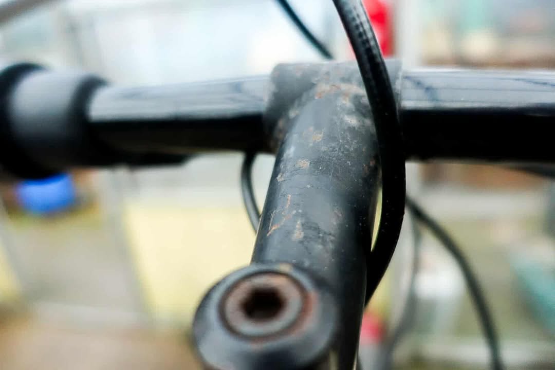 Close-up of a rusty, black metal handlebar on a bicycle, with visible rust around the stem, against a blurred background.