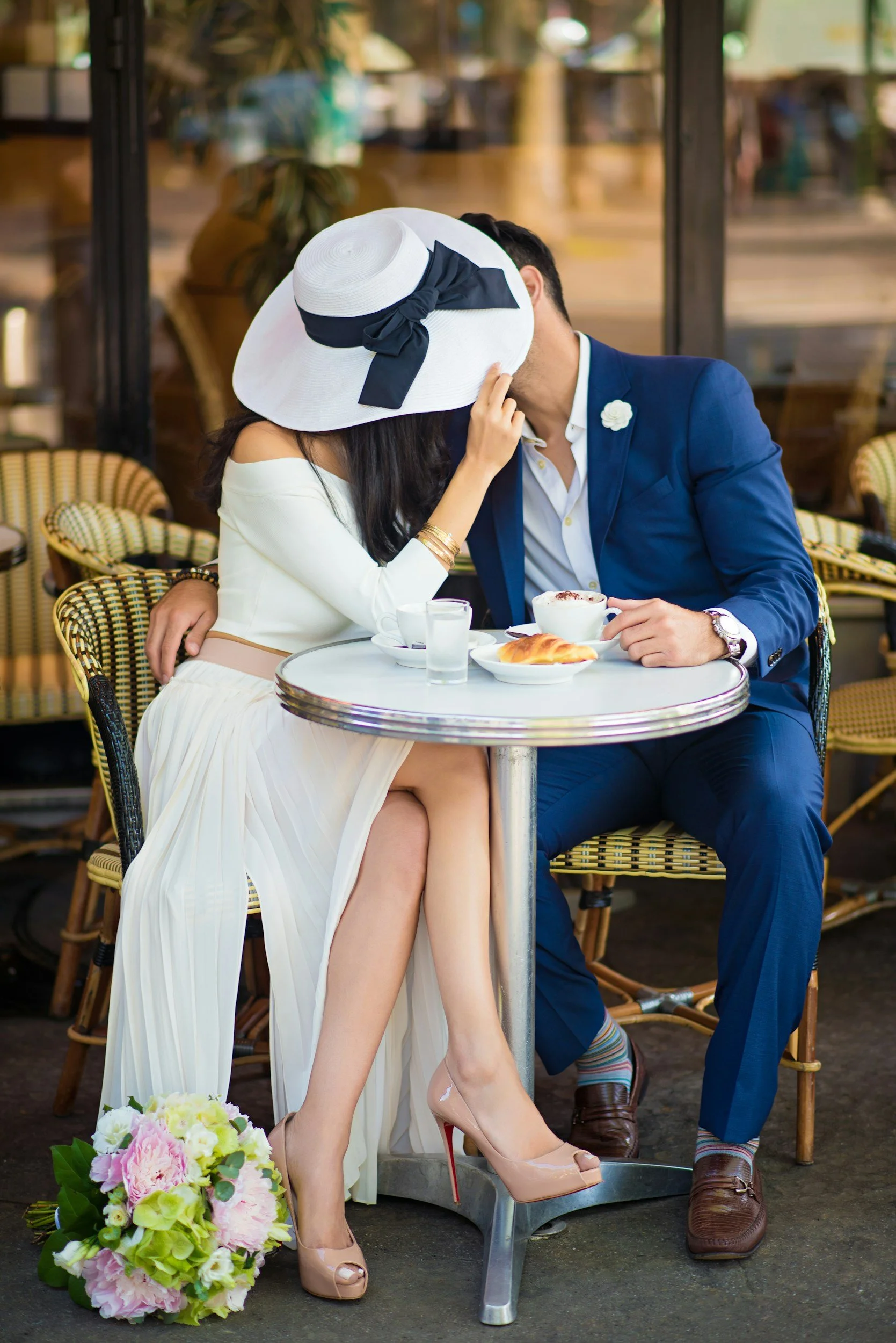 A couple sitting closely at a café table, sharing a kiss, with a woman wearing a large white sunhat with a black ribbon and a white dress, and a man in a blue suit and white shirt, with dessert and coffee on the table, and flowers on the ground nearby.