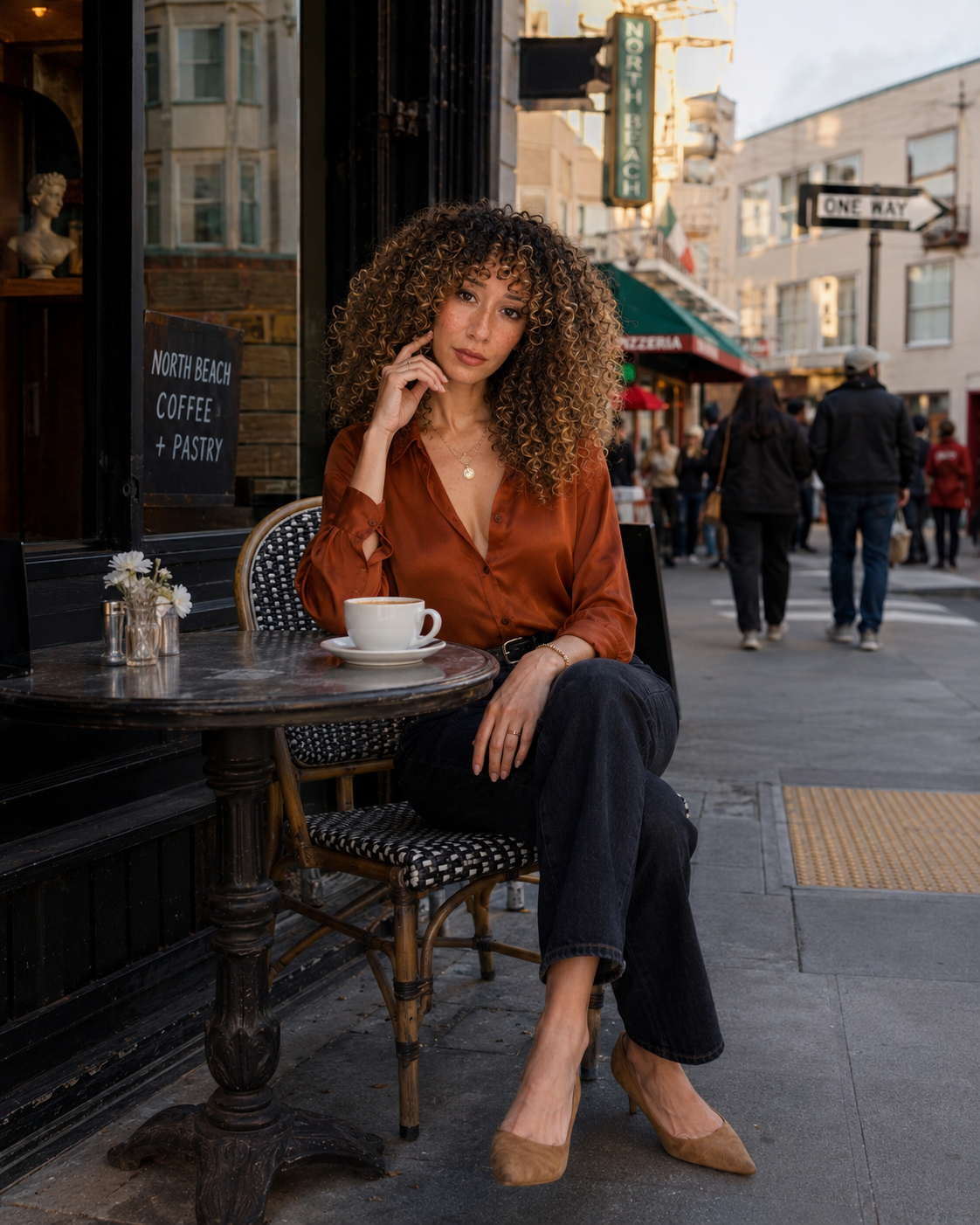 A woman with curly hair and freckles sitting outside a café on a city street, holding a cup of coffee. She is wearing a rust-colored shirt and black pants, with a small flower arrangement on the table. The background shows pedestrians and storefronts, with signs indicating north beach and one way.