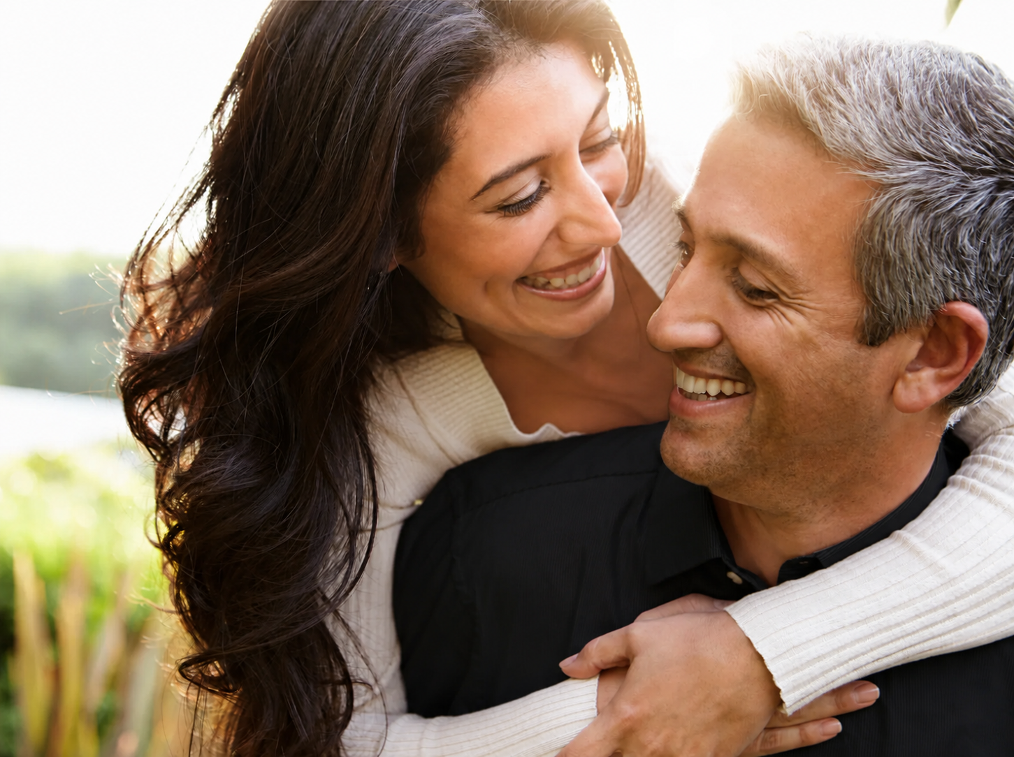 A woman with long dark hair and a man with short gray hair smiling and embracing each other outdoors in sunlight.