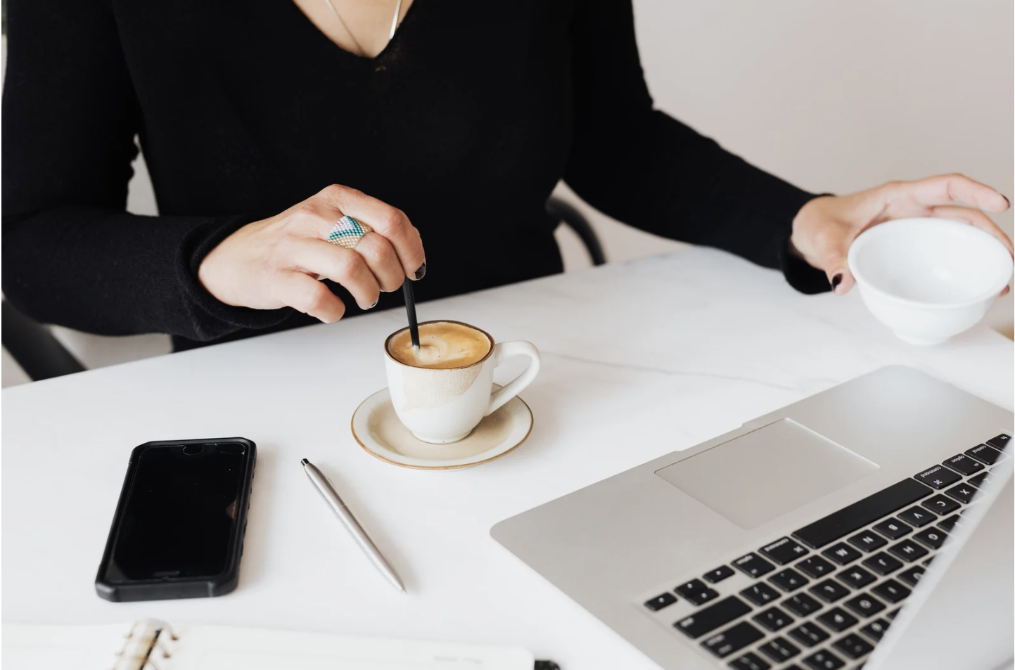 A person in a black long-sleeve shirt stirring a cup of coffee with a black straw, holding an empty white bowl, with a laptop, smartphone, pen, and notebook on a white desk.